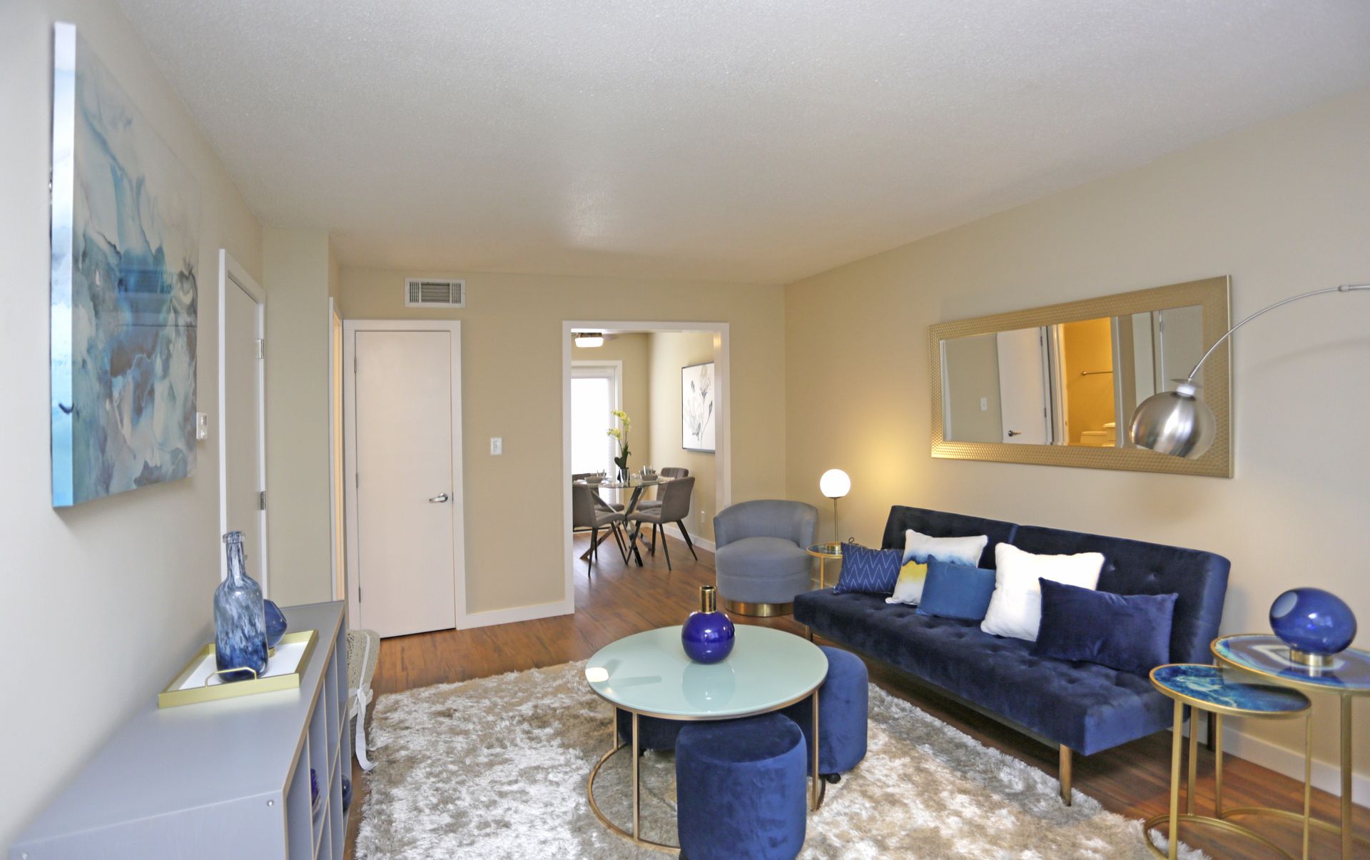 Living room with blue sofa, white rug, and gold accents. Beige walls.