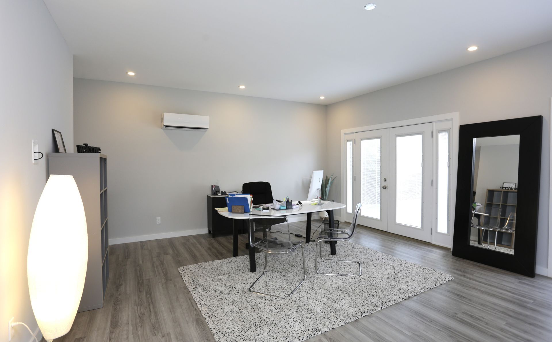 Modern office with gray walls, desk, and rug. French doors, floor lamp, and full-length mirror.