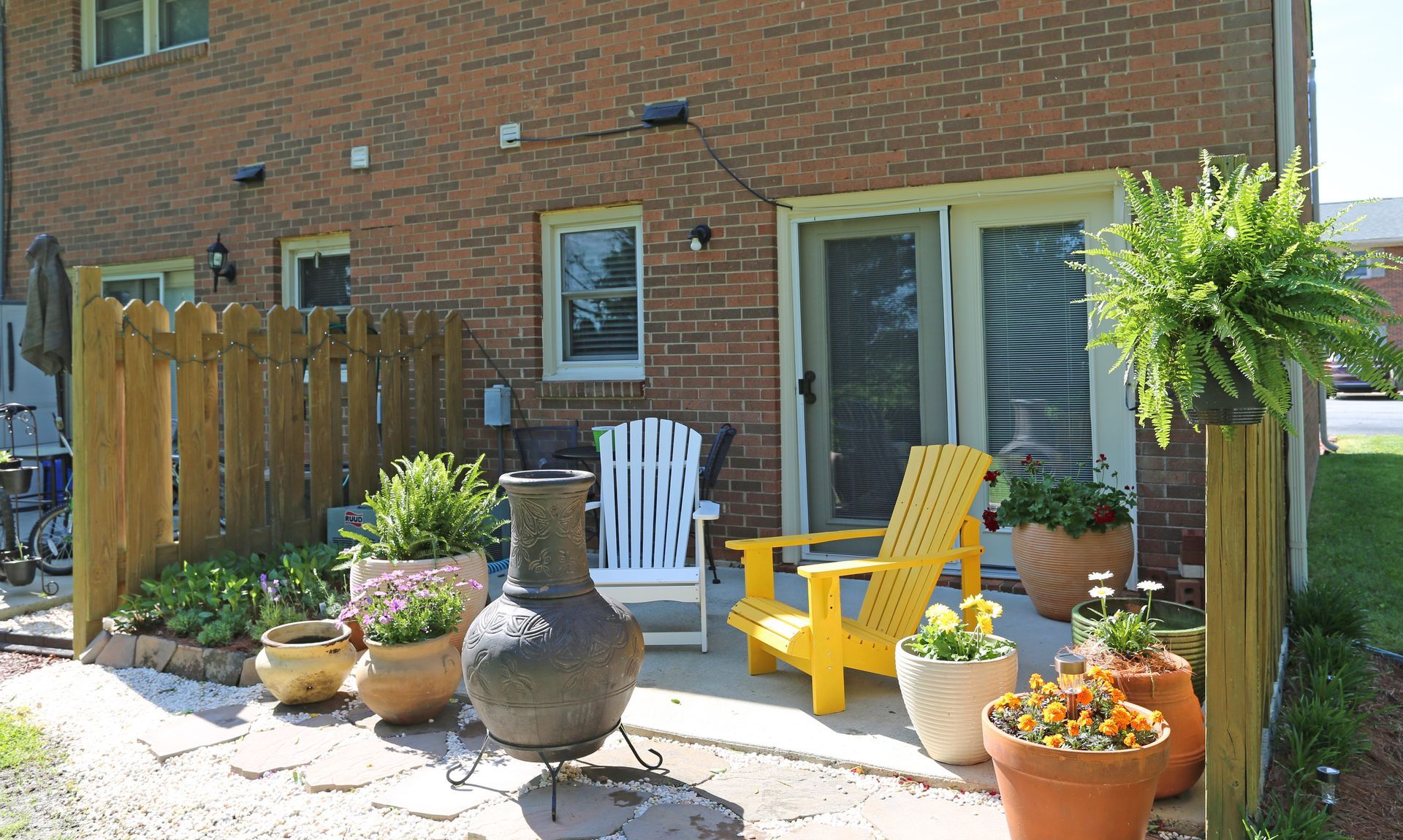 Patio with brick building backdrop; wooden fence, chairs, potted plants, and a fern.