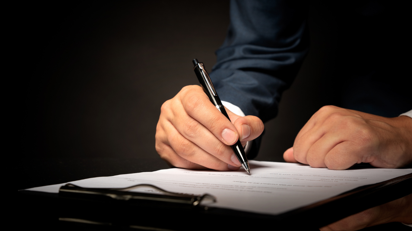 Hands signing a document with a pen at a desk under low light.