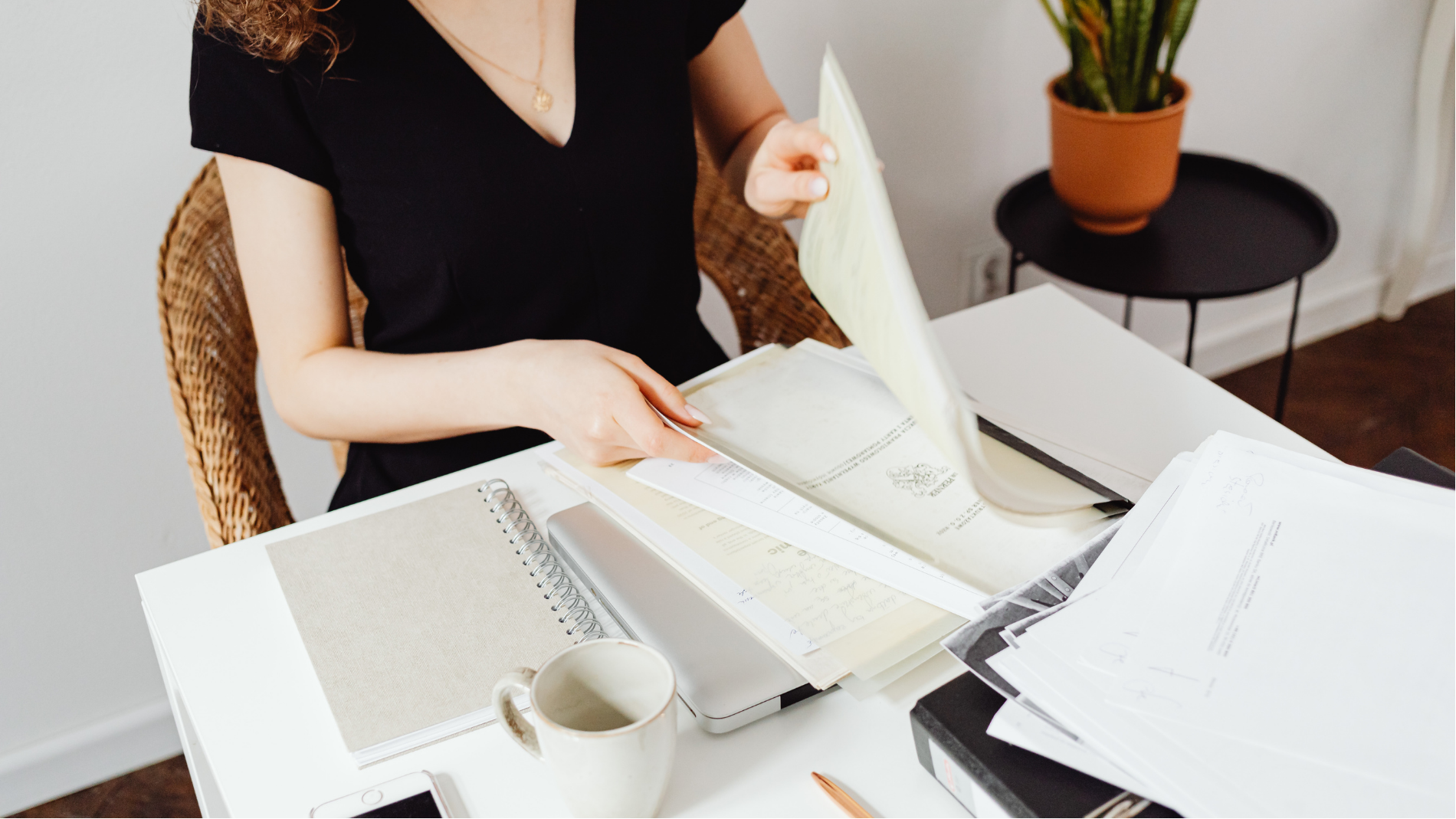 Person at a desk reviewing papers with a coffee mug and a potted plant nearby