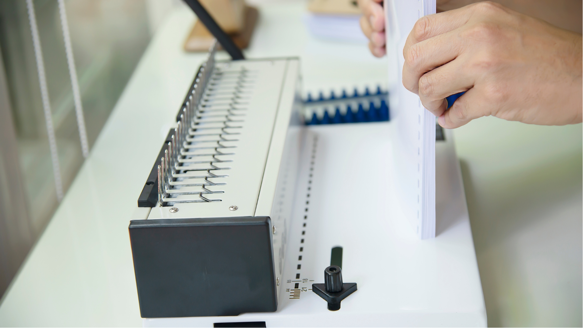 Hands assembling a white spiral-bound notebook on a workbench with metal binding machine