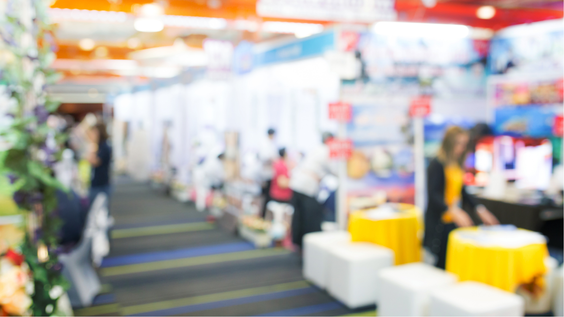 Blurred trade show booth aisle with people, colorful displays, and white cube seating on the right