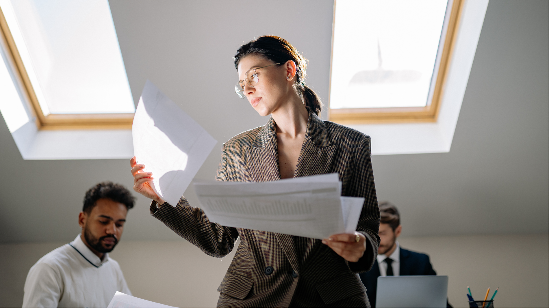 Office team reviewing documents under skylight, with one person standing and holding papers while others work nearby