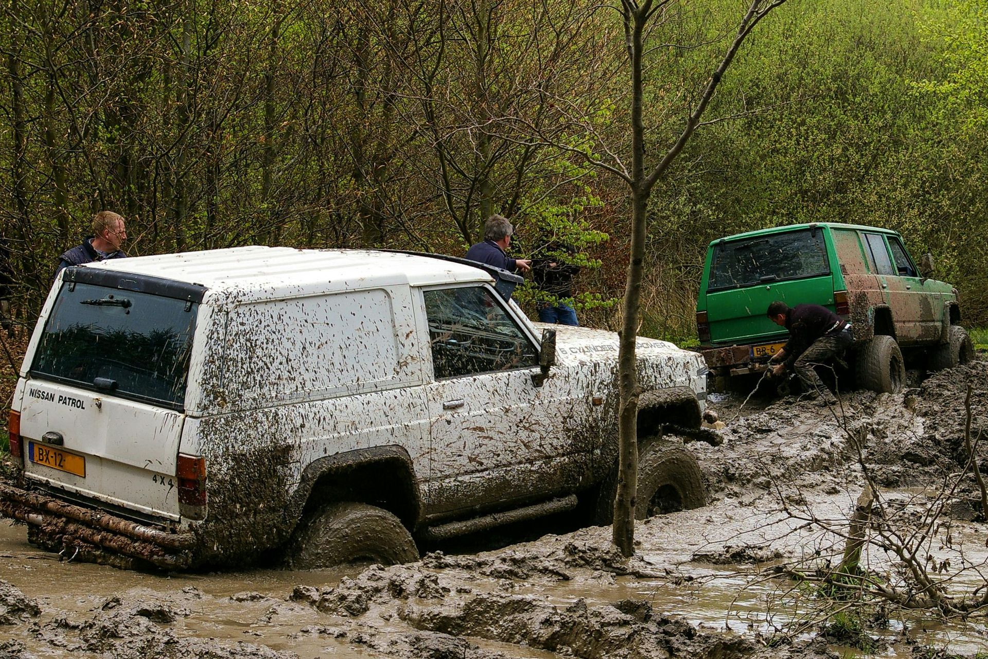 Two muddy off-road vehicles stuck in a mud pit, two people appear to be helping, one wearing green.