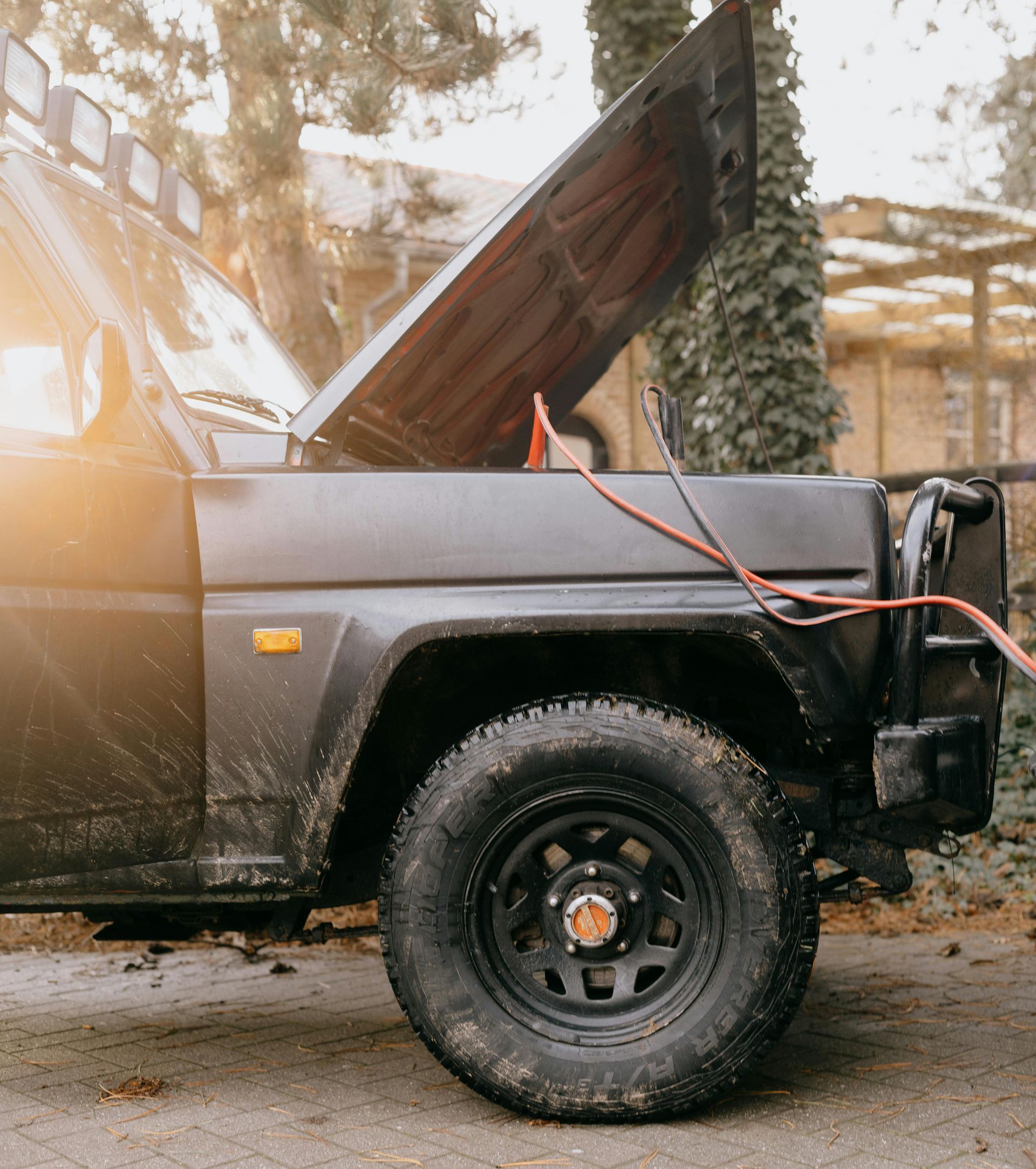 Black SUV with open hood being jump-started. Muddy tire in foreground, cables attached. Outdoors.
