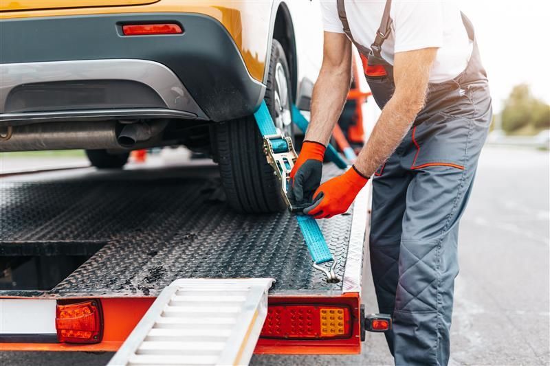 A tow truck operator securing a yellow car with blue straps on a flatbed trailer.