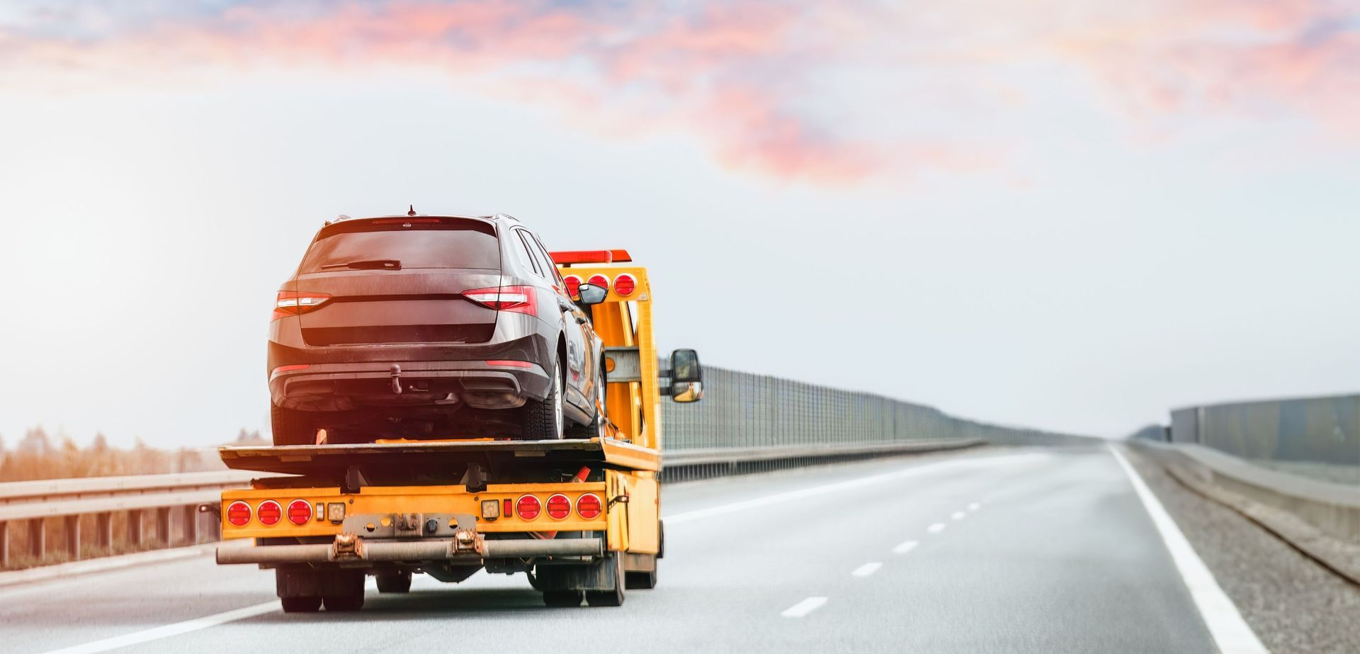 A black SUV being towed on a highway by a yellow tow truck. Overcast sky.