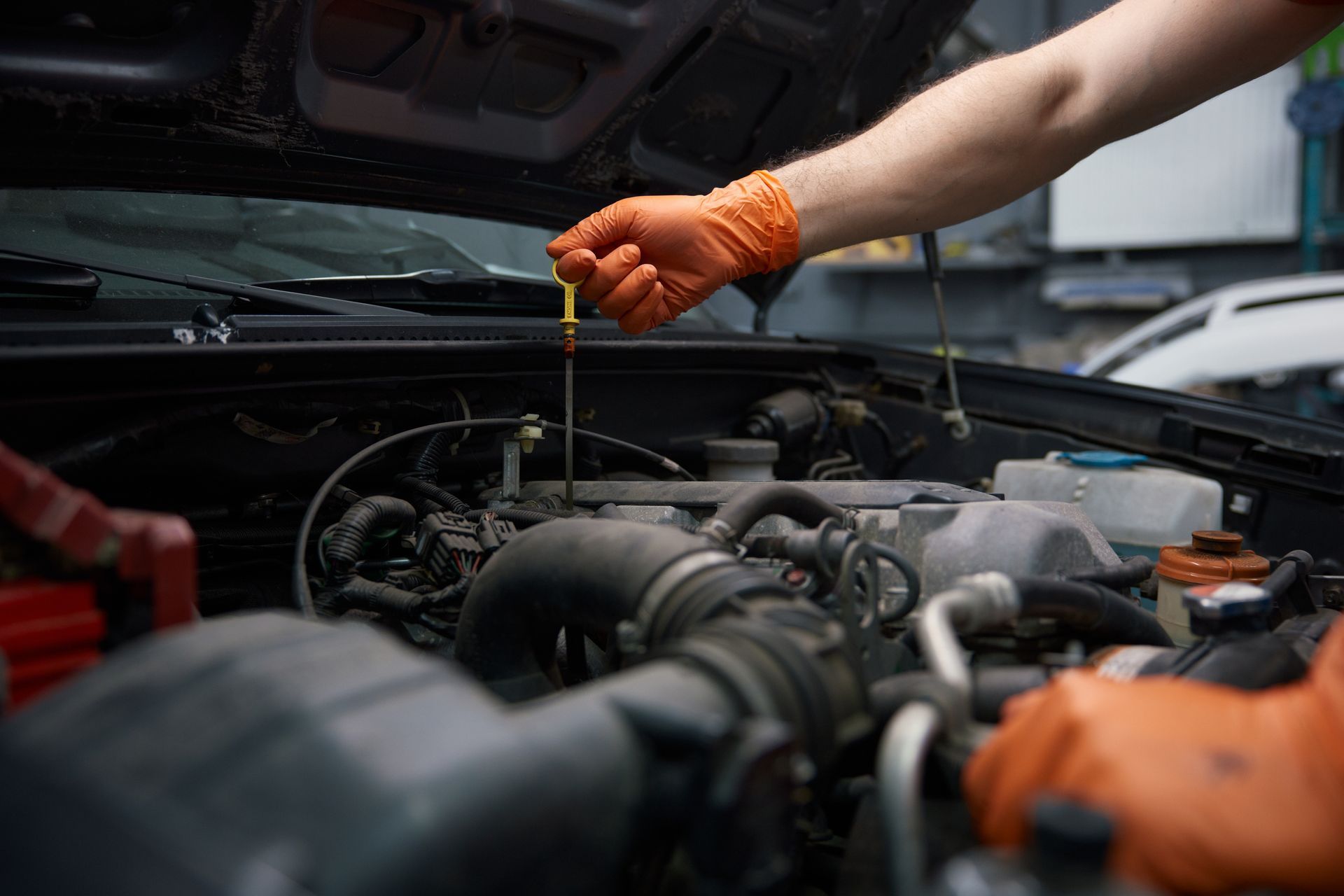 Mechanic checking engine oil with a dipstick, wearing orange gloves in an open car hood