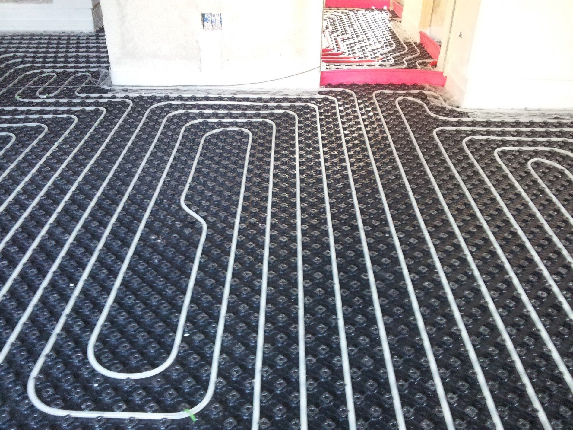 Tubi in plastica bianca per il riscaldamento a pavimento radiante, installati a griglia su pannelli isolanti neri a rilievo.