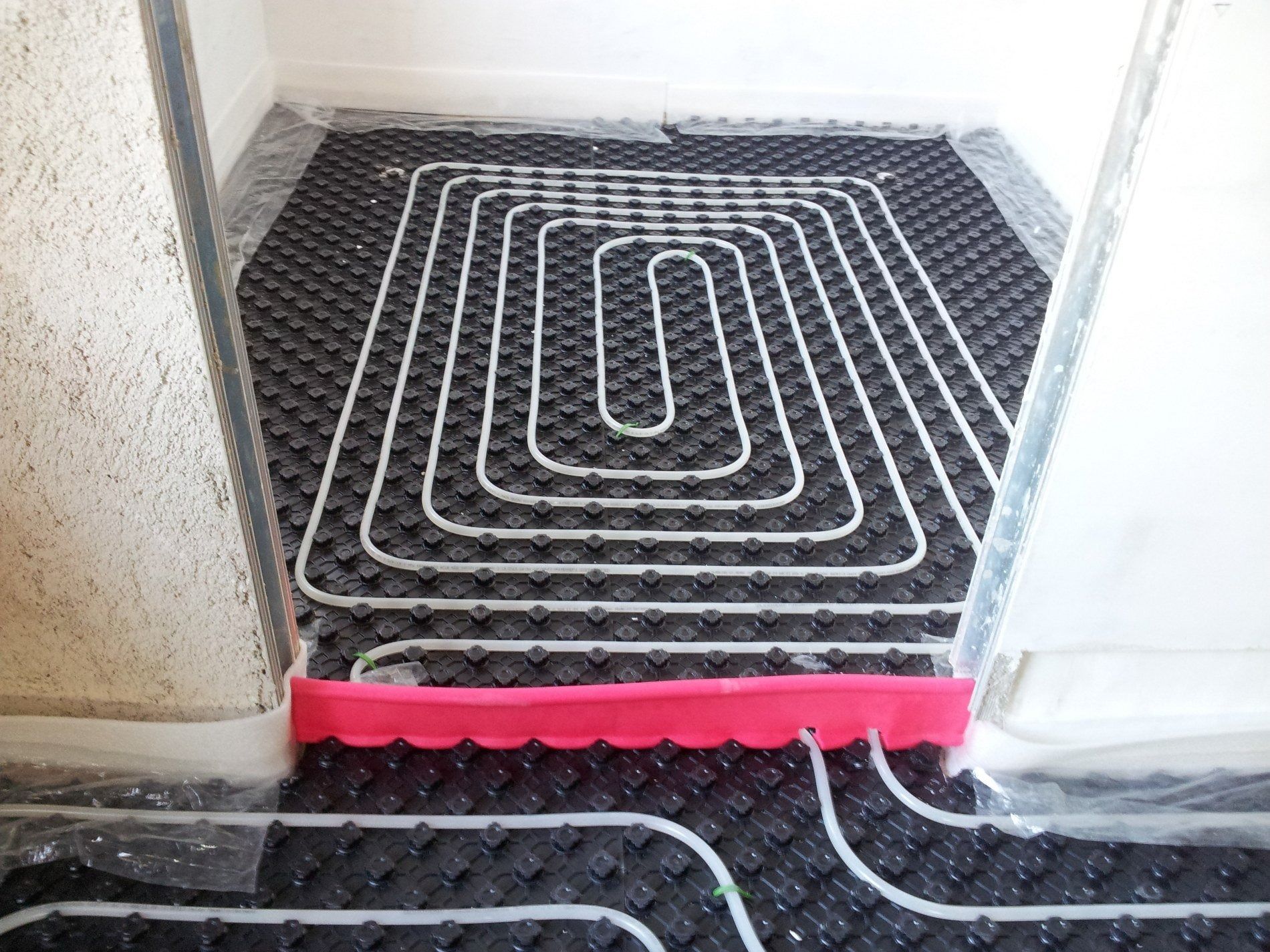 Tubi bianchi per il riscaldamento a pavimento, disposti a spirale su pannelli isolanti neri, con una striscia di confine rossa.