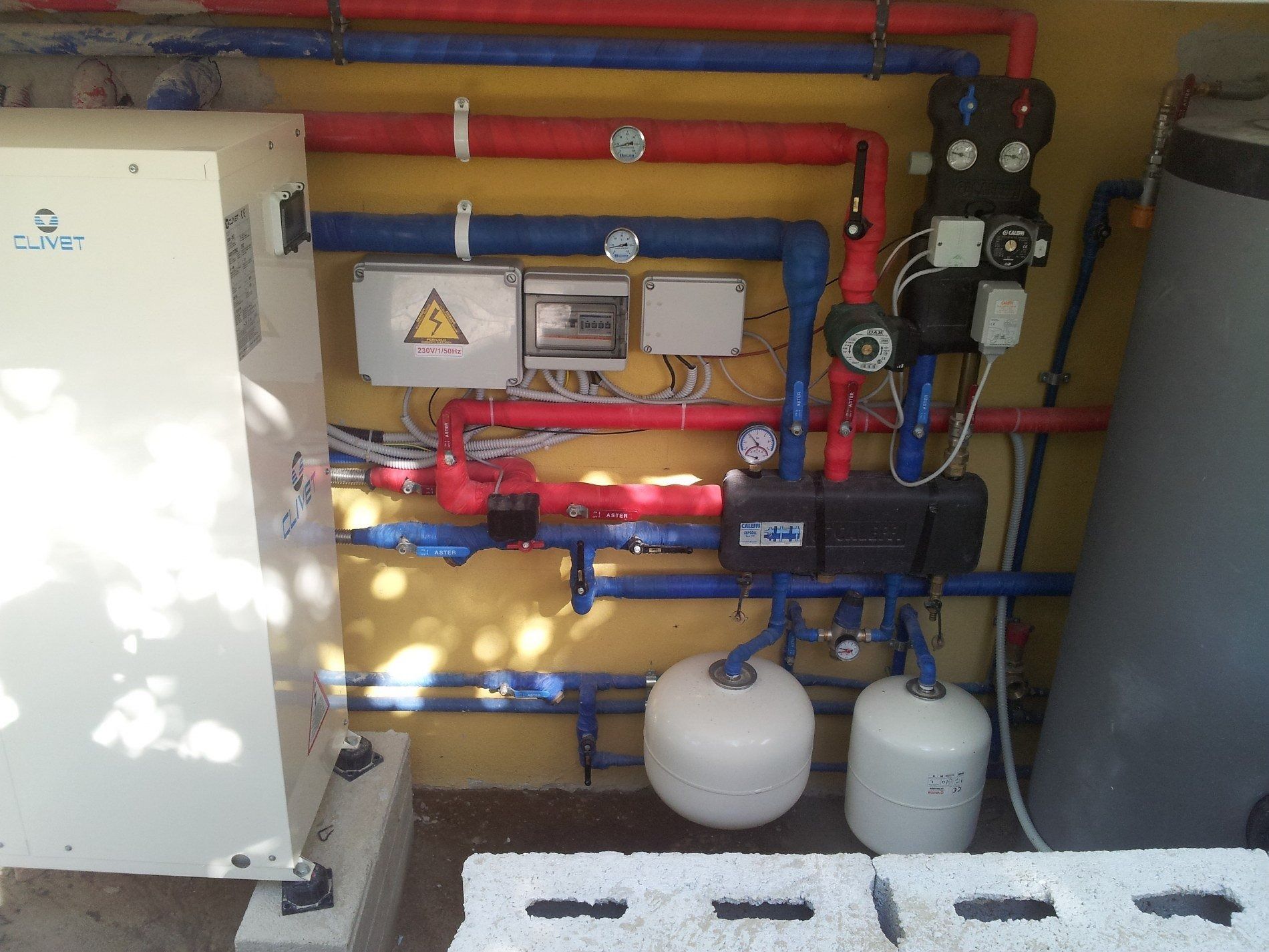 Un sistema di pompa di calore residenziale con tubazioni rosse e blu, quadri elettrici di controllo e due vasi di espansione bianchi.