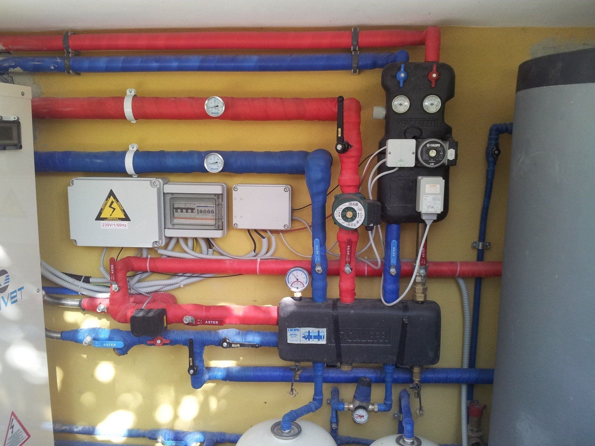 Un complesso sistema idraulico e di controllo HVAC con tubi isolati rossi e blu, scatole elettriche e pompe su una parete gialla.