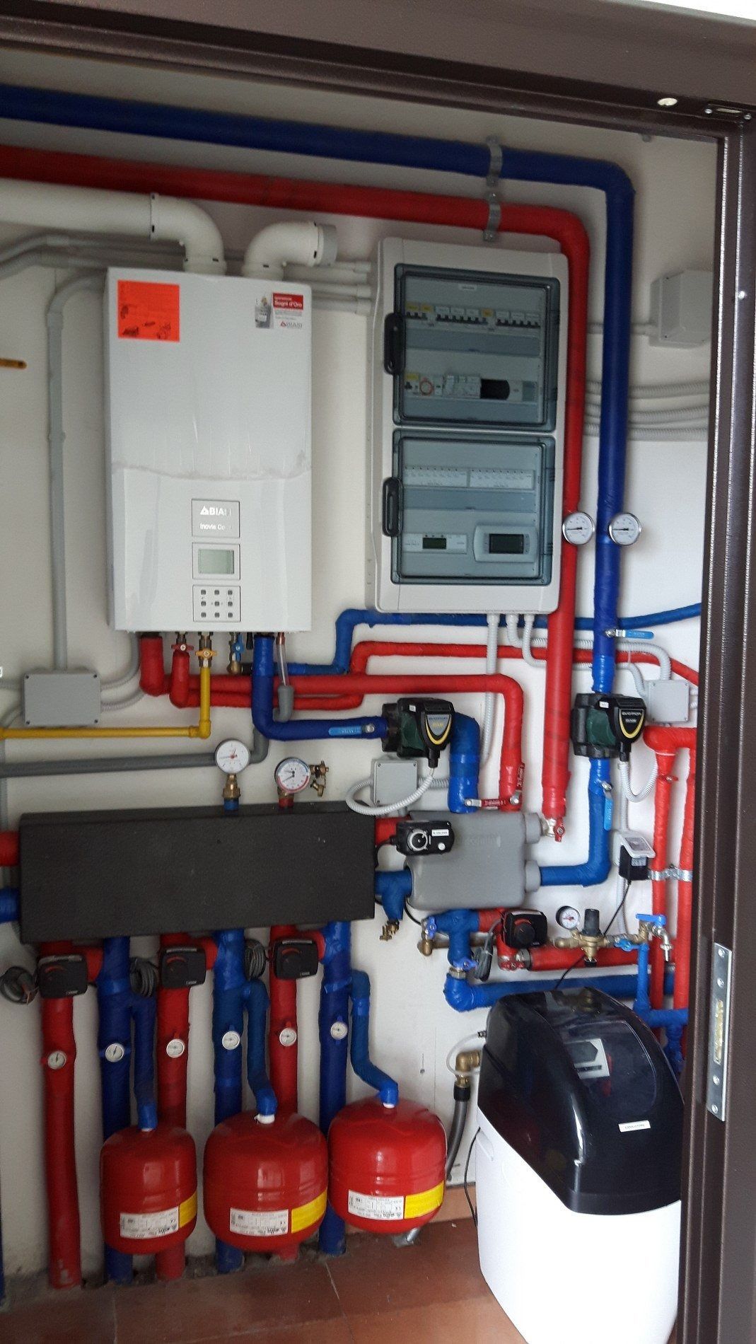 Unità centrale di riscaldamento e caldaia per uso residenziale, con tubi, manometri e vaso di espansione rosso, montata su una parete bianca.