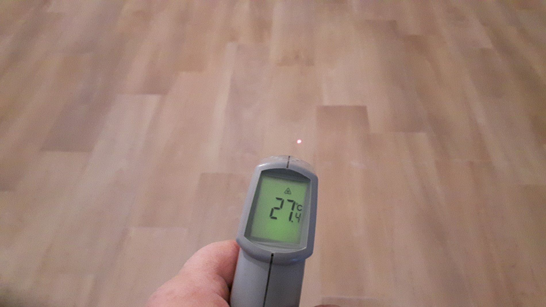 Una mano che impugna un termometro a infrarossi puntato verso un pavimento effetto legno, con il display digitale che mostra 27 gradi Celsius.
