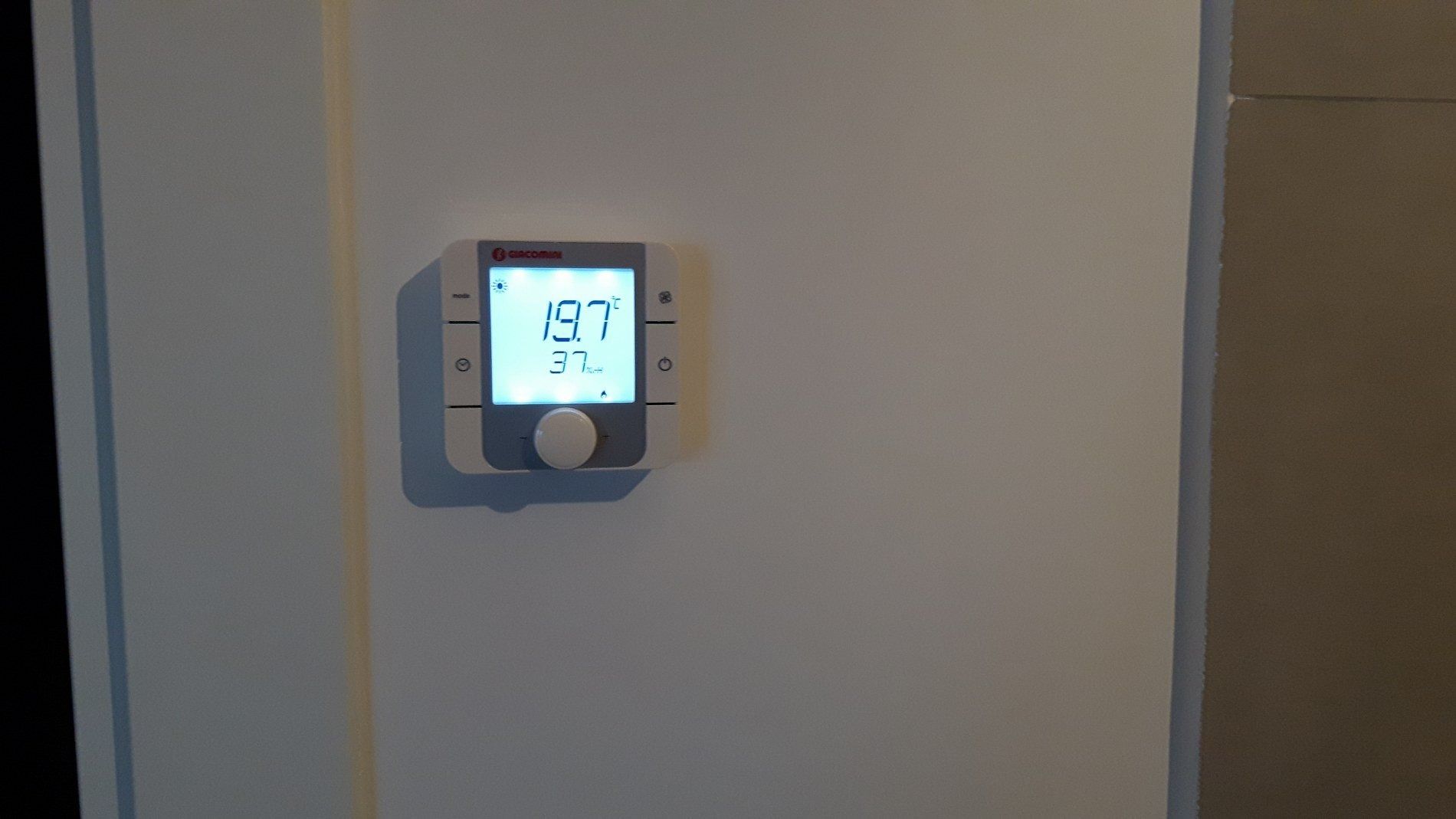 Un termostato digitale montato su una parete bianca visualizza una temperatura di 19,7 gradi Celsius.
