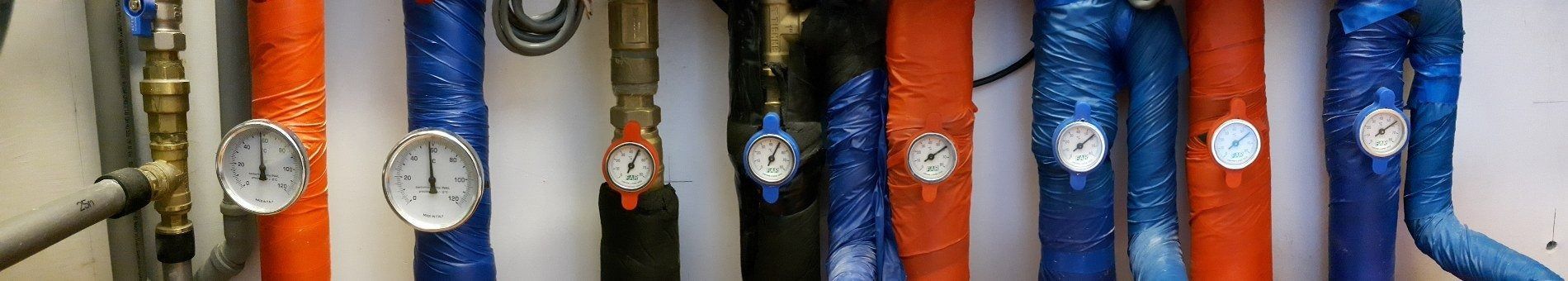 Una fila di tubi industriali con guaine isolanti arancioni e blu, ciascuno dotato di un manometro circolare.