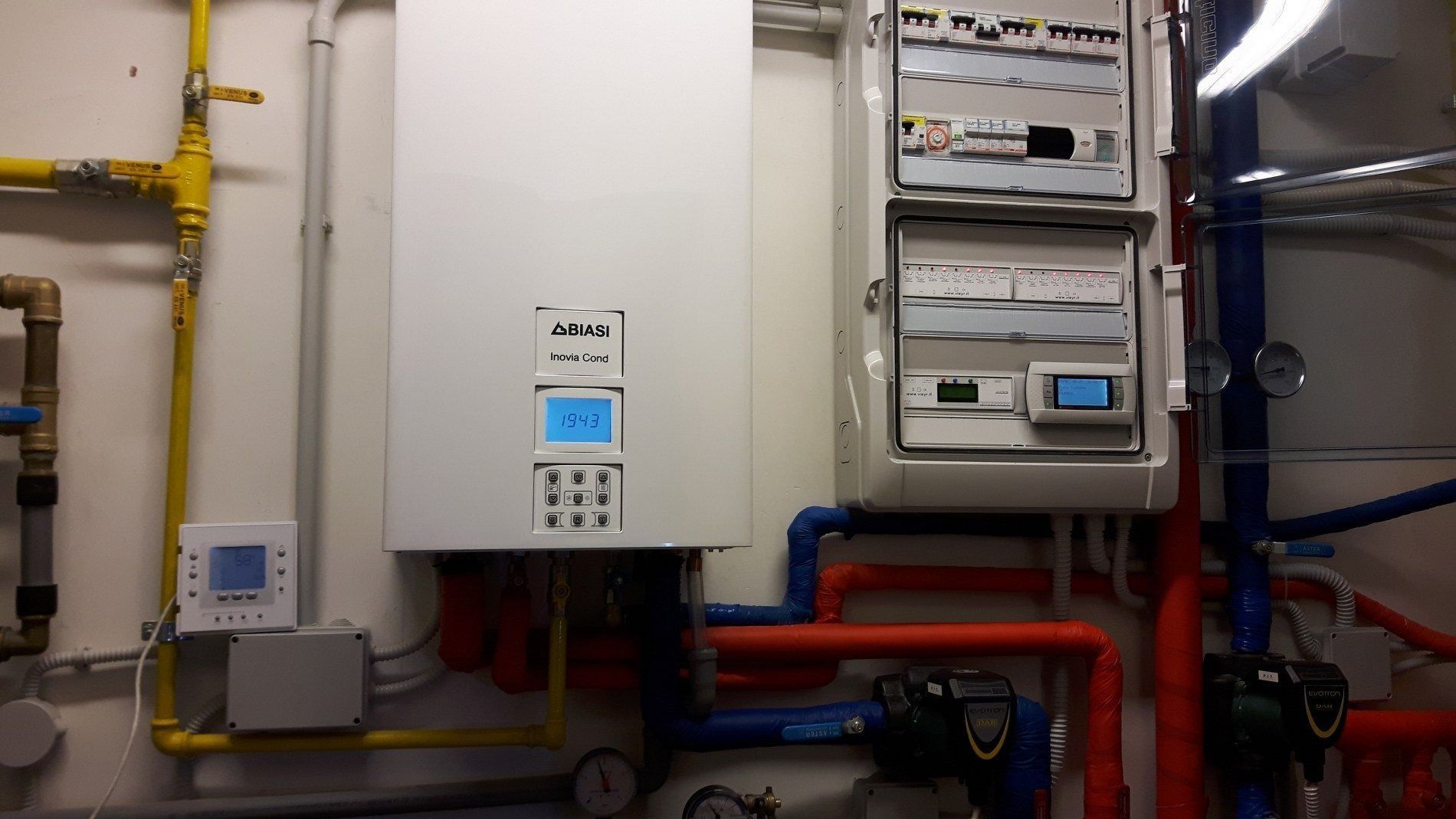 Caldaia a parete e quadro elettrico di controllo con vari tubi, indicatori e termostato digitale in un locale di servizio.