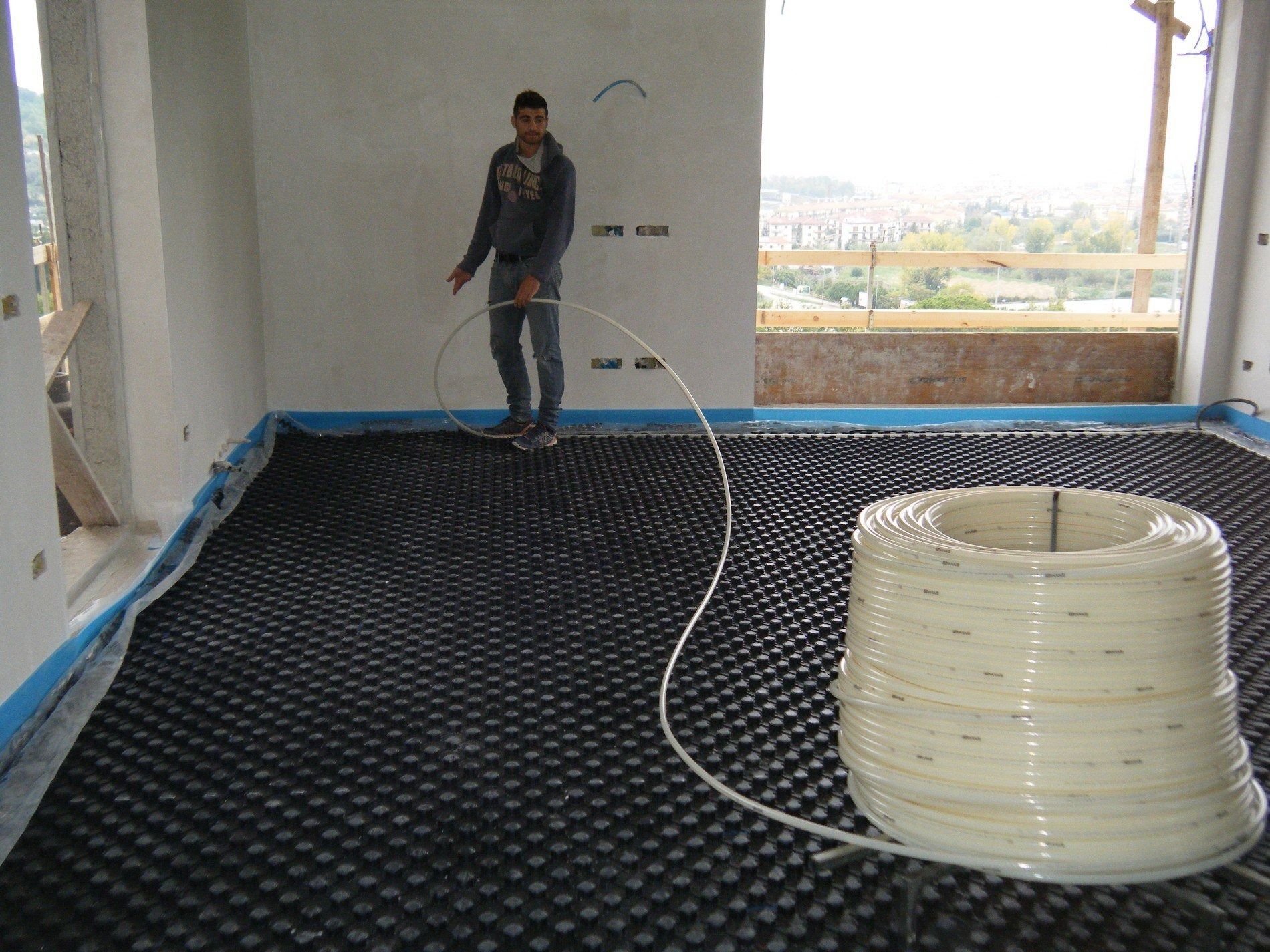 Un operaio installa tubi flessibili in plastica per il riscaldamento a pavimento radiante su una griglia di pannelli sagomati neri in una stanza non arredata.