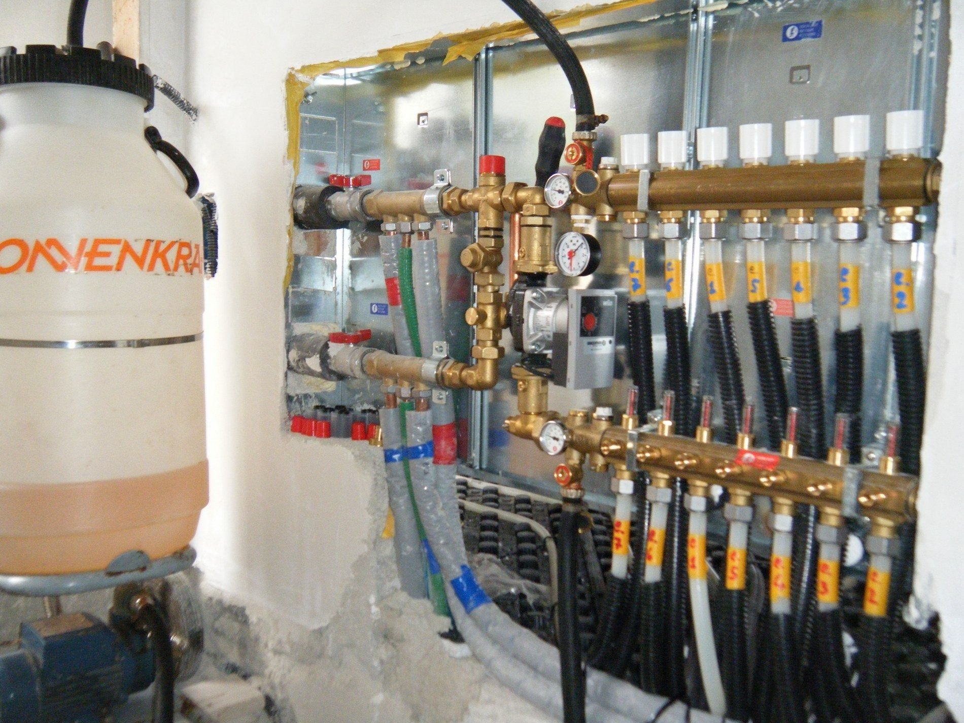 Sistema di riscaldamento radiante idronico a parete con collettore, tubi multipli, manometri e un grande serbatoio sulla sinistra.