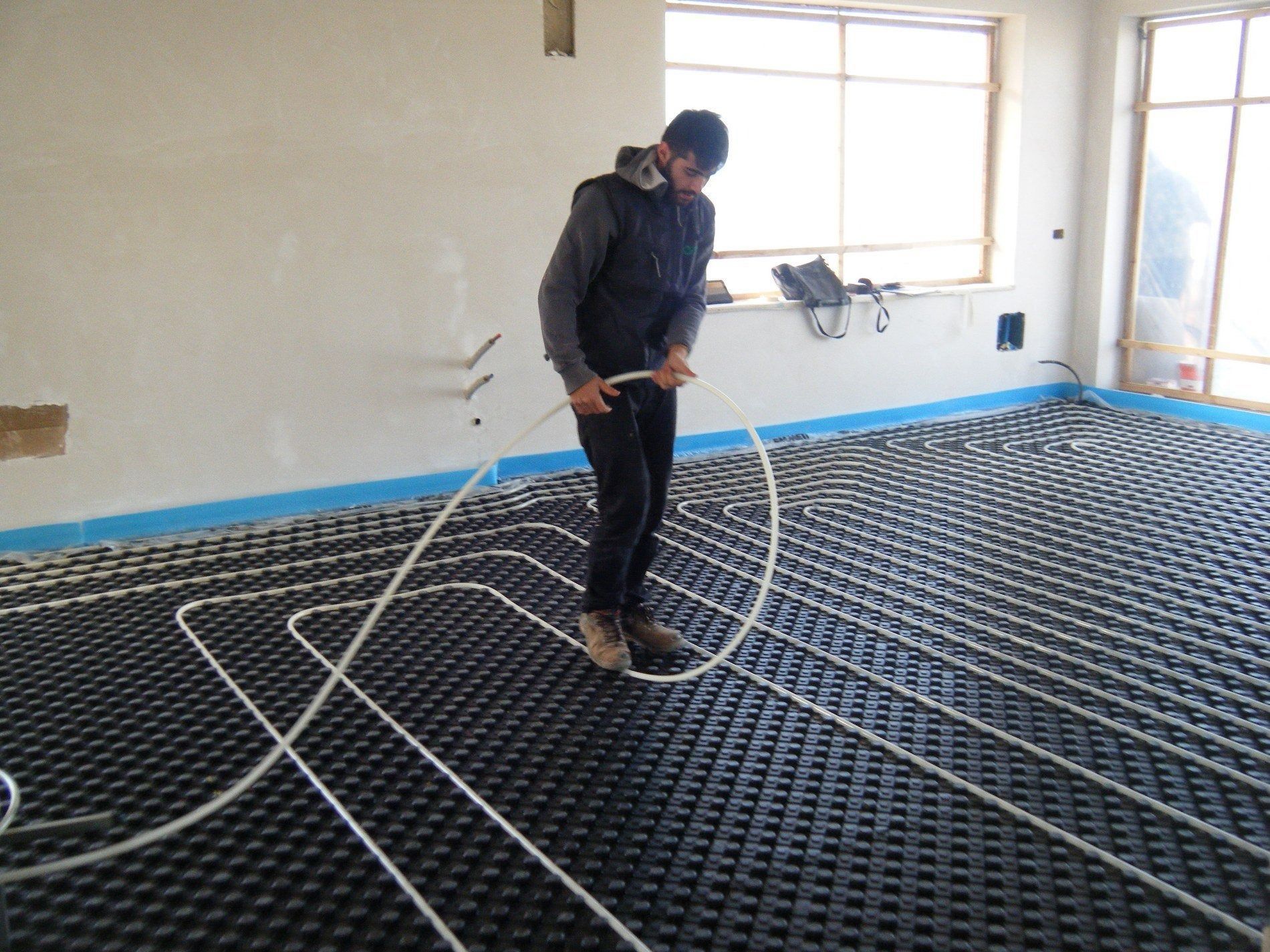 In una stanza in costruzione, una persona installa dei tubi bianchi per un sistema di riscaldamento a pavimento radiante su pannelli a griglia neri.