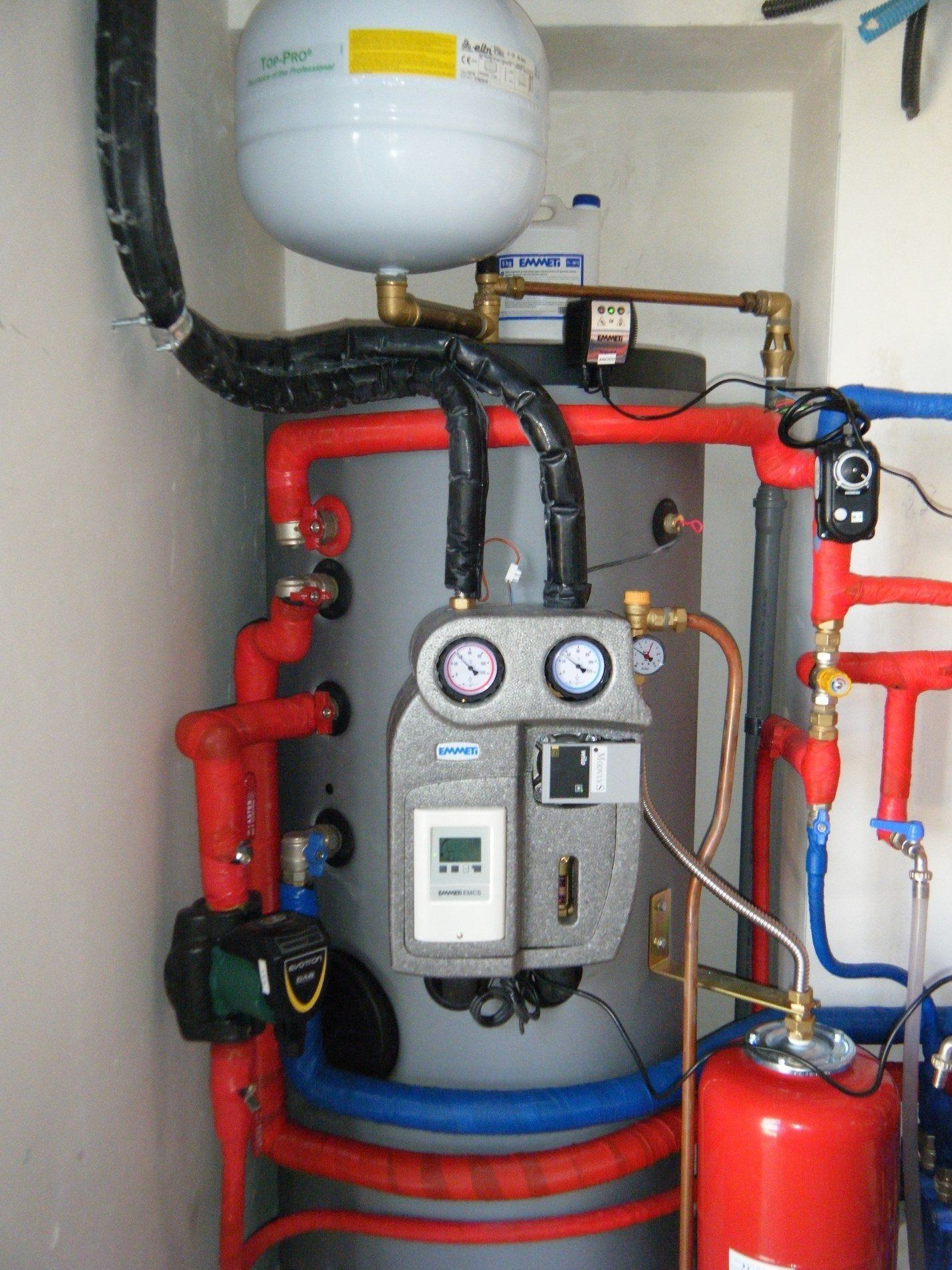 Un sistema di caldaia grigio con tubi isolati rossi e blu, due manometri e un regolatore digitale in un locale tecnico.