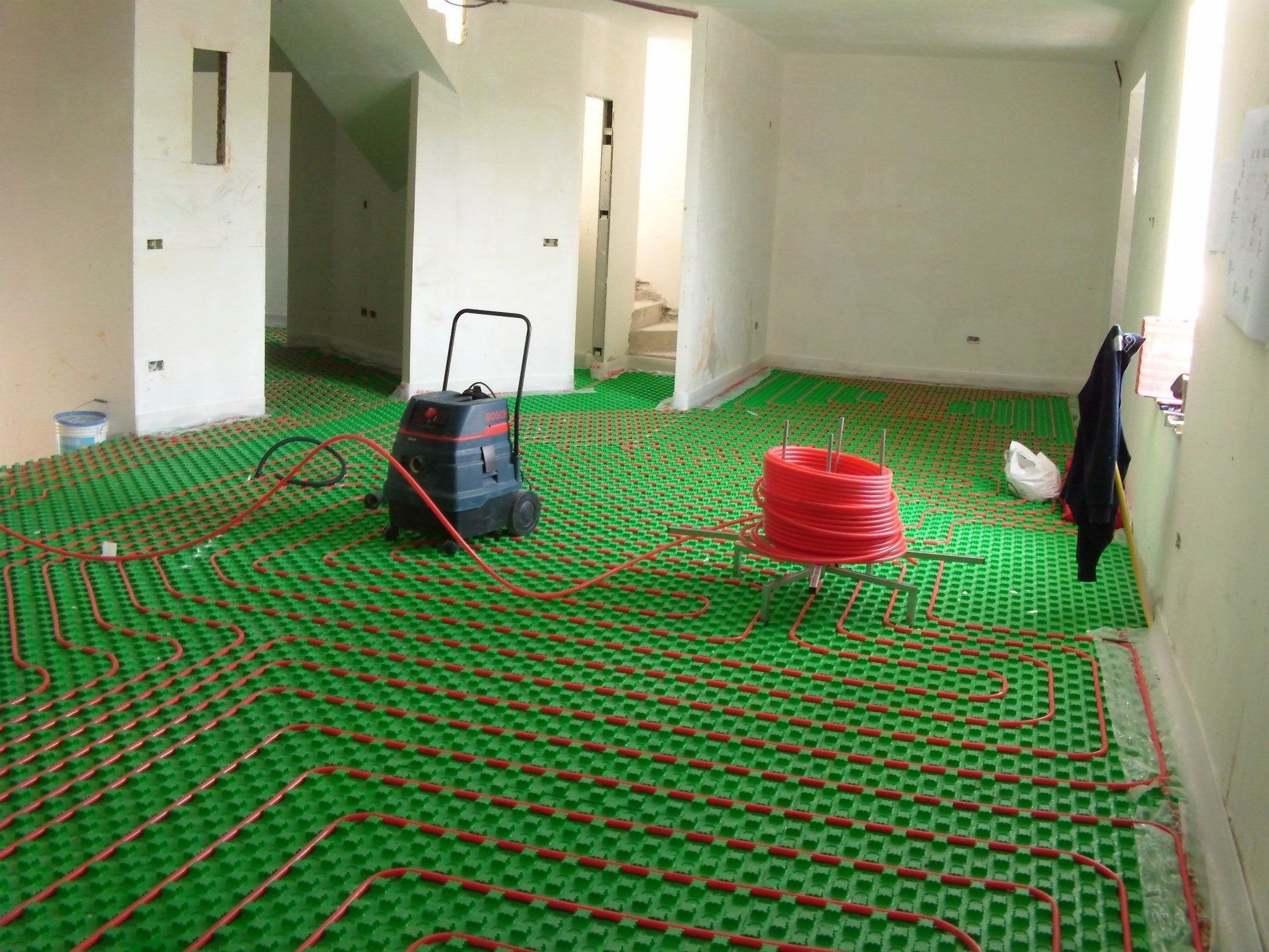 Una stanza in costruzione con tubi per il riscaldamento a pavimento radiante installati su una base a griglia verde, con le apparecchiature nelle vicinanze.