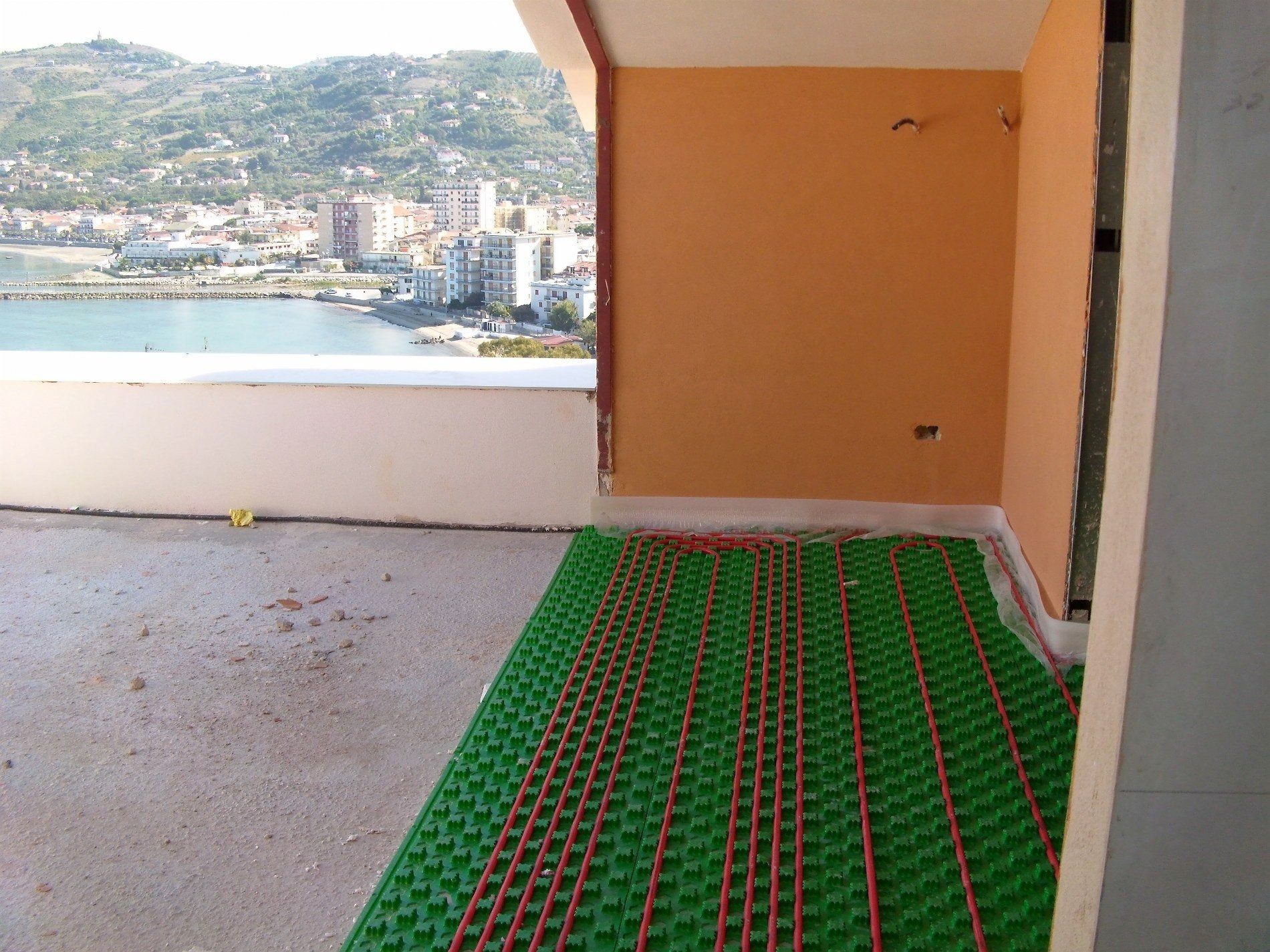 Tubature per il riscaldamento a pavimento installate su pannelli isolanti verdi in una stanza con vista su una cittadina costiera e sul mare.