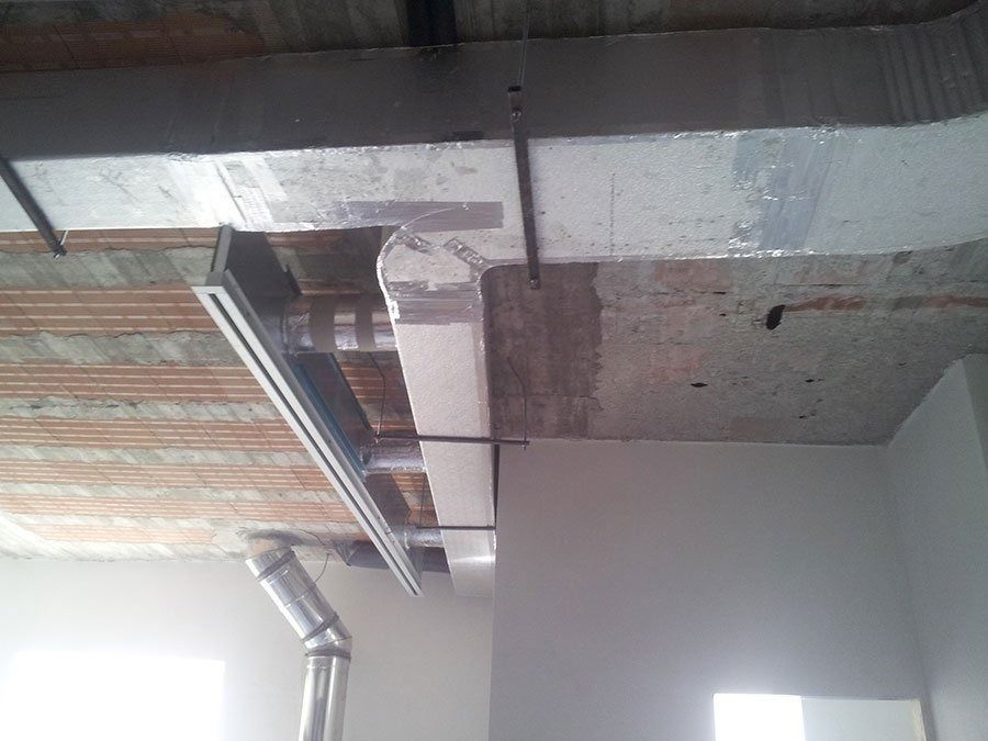 Vista di un soffitto con canalizzazioni per impianti di climatizzazione, tra cui una griglia di ventilazione metallica, tubi isolati e supporti a sospensione, all'interno di una stanza.