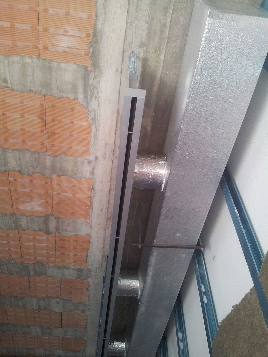 Una lunga griglia di ventilazione industriale color argento, montata su una parete tra una sezione in mattoni e un pannello bianco testurizzato.