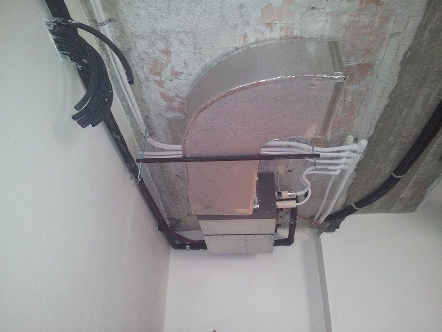 Un condotto di ventilazione metallico rettangolare installato su un soffitto in cemento, con cavi neri a vista e tubature bianche.
