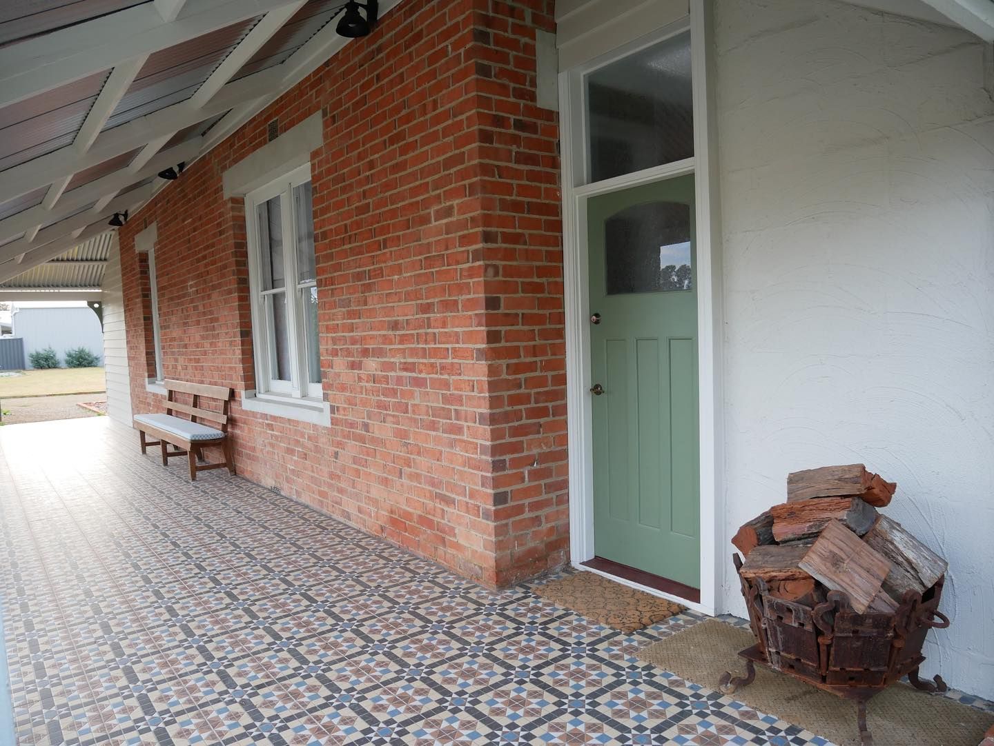 Brick veranda with patterned tile floor and green door. A bench and wood pile are visible.