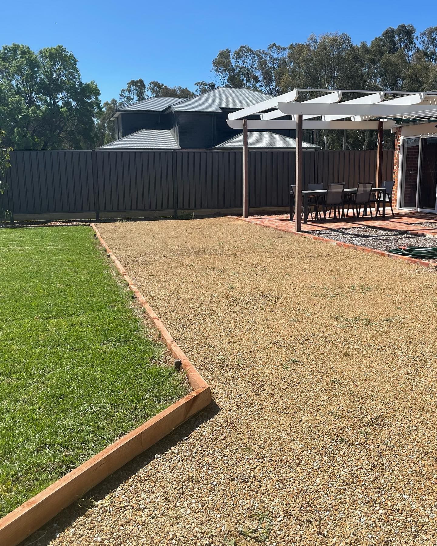 A backyard with gravel, green grass, a pergola, and a wooden fence under a blue sky.