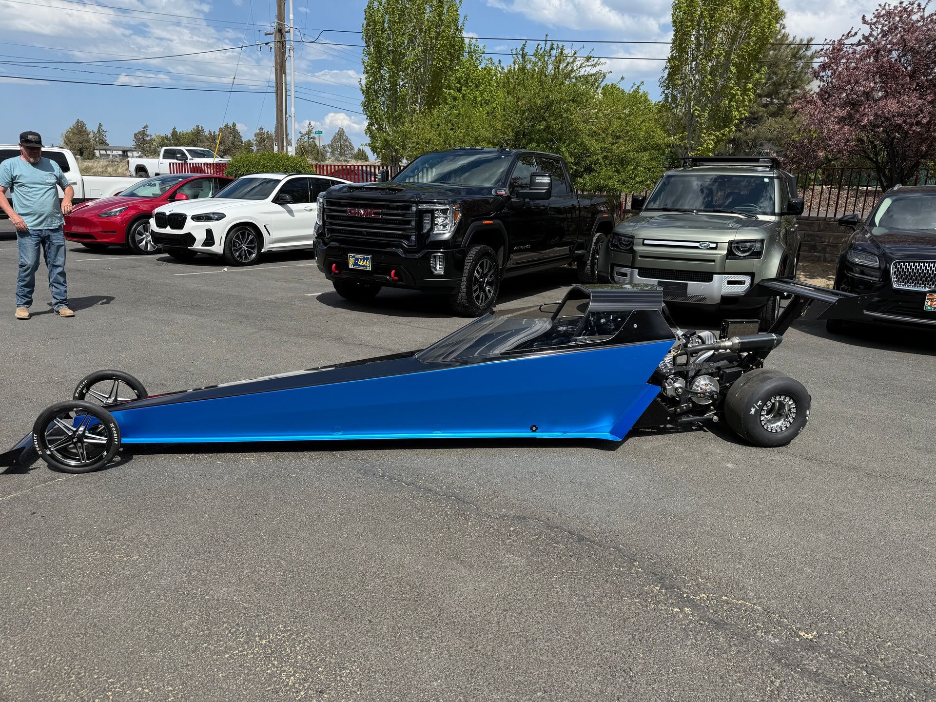 A man is standing next to a blue dragster in a parking lot.