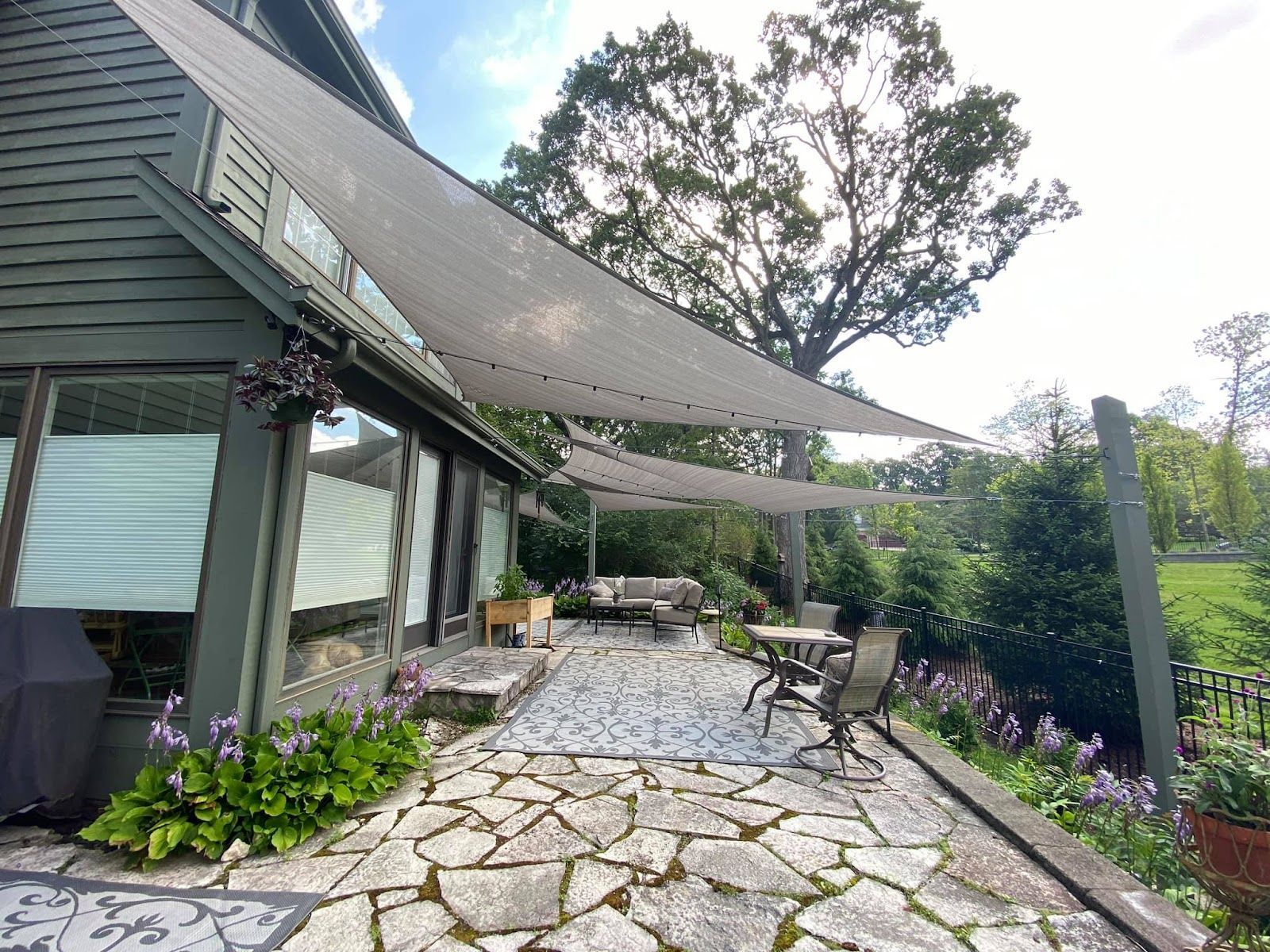 Patio with stone pavers, seating, and shade sails, backed by a house and lush greenery.