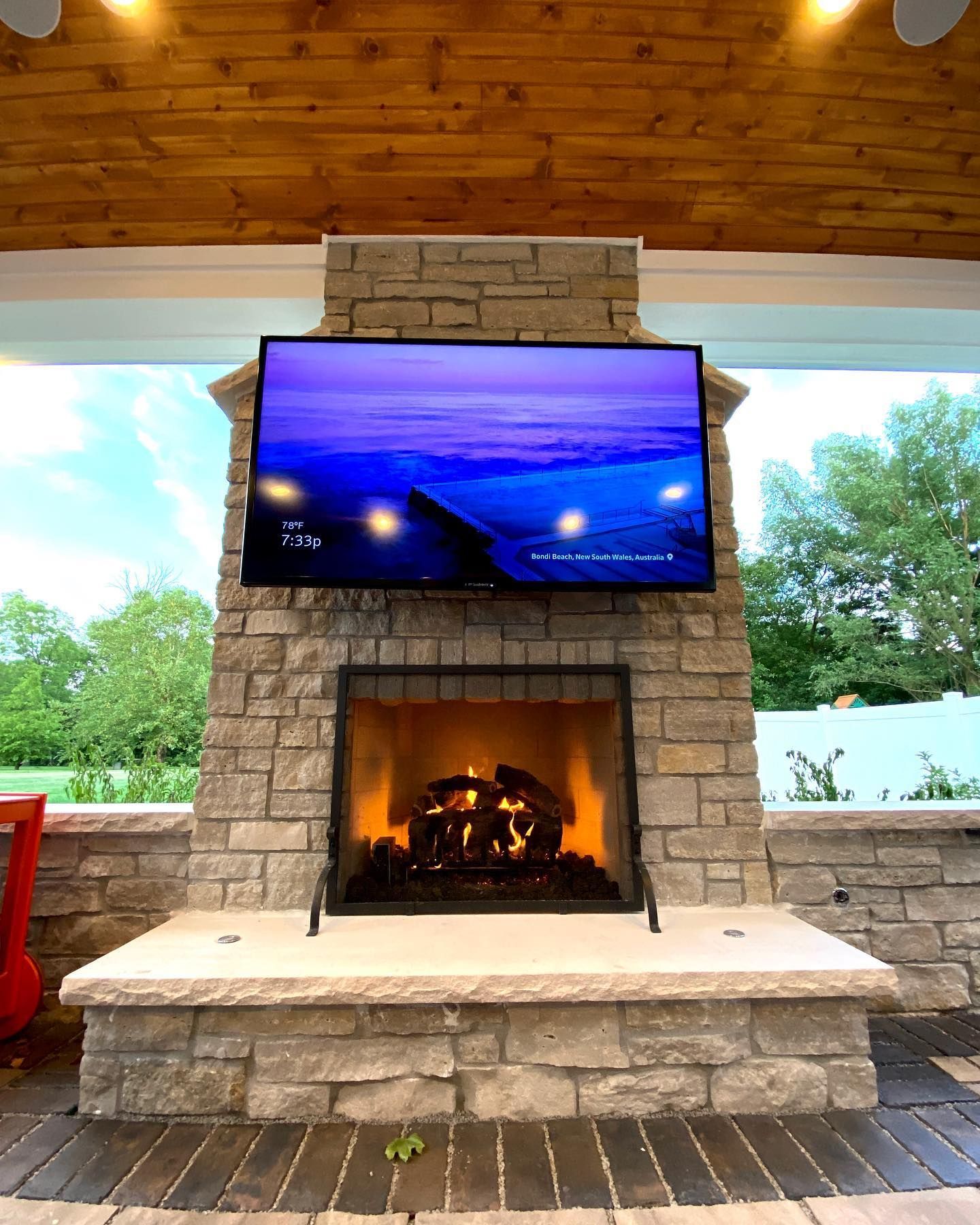 Outdoor fireplace with a lit fire, stone facade, and a mounted TV displaying a blue image, under a wooden ceiling.