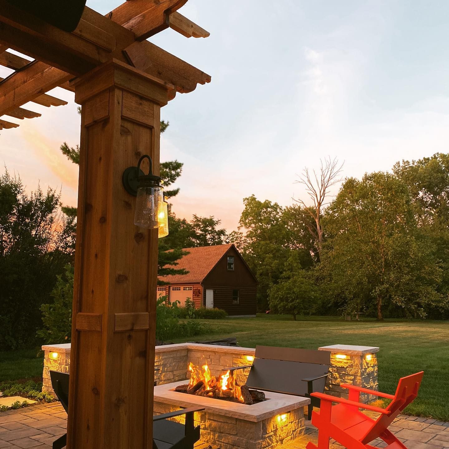 Outdoor patio with fire pit, wooden pergola, and orange Adirondack chair in a grassy yard at dusk.