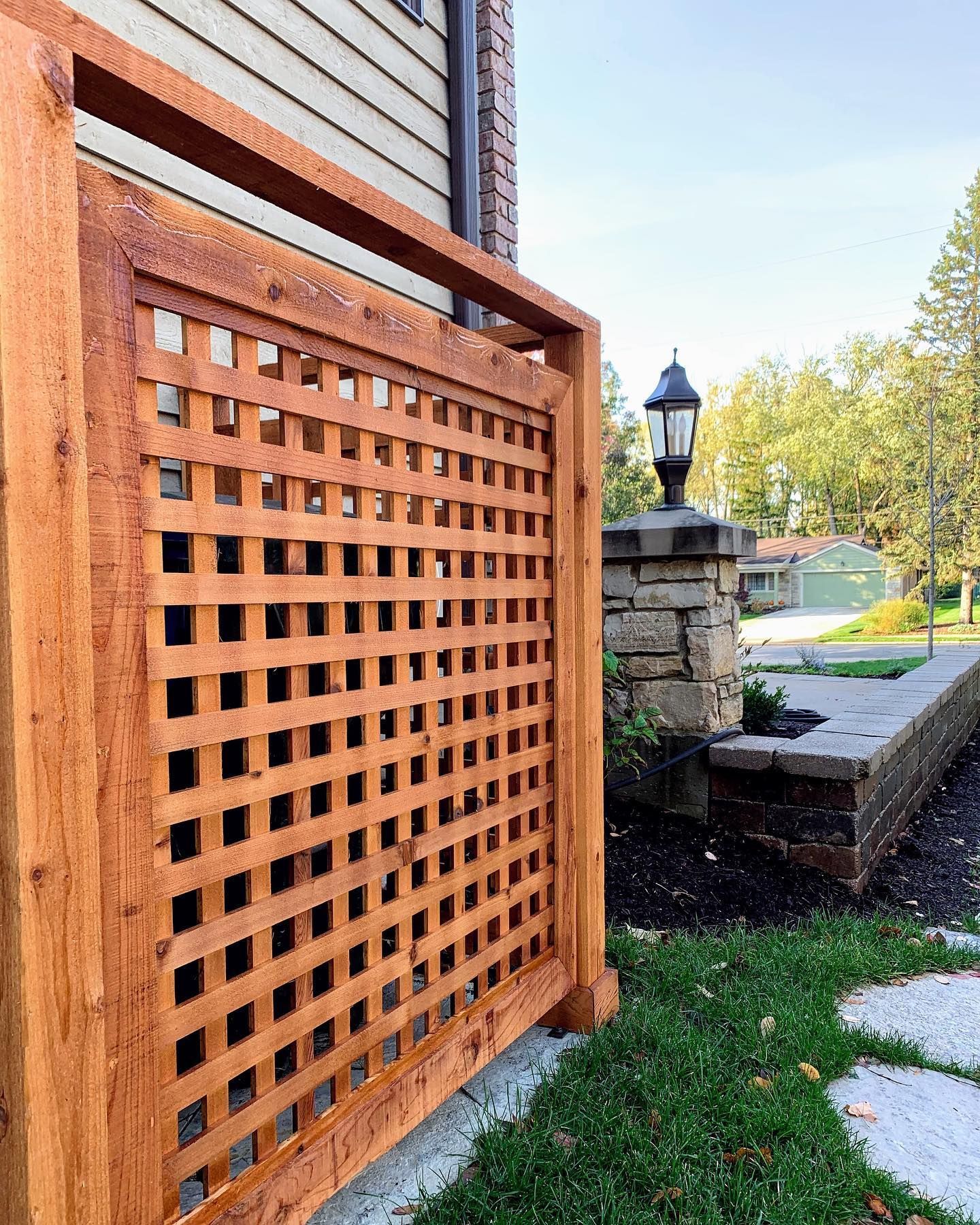 Wooden lattice privacy screen near a brick and stone pillar with a lamp, set on a grassy area.