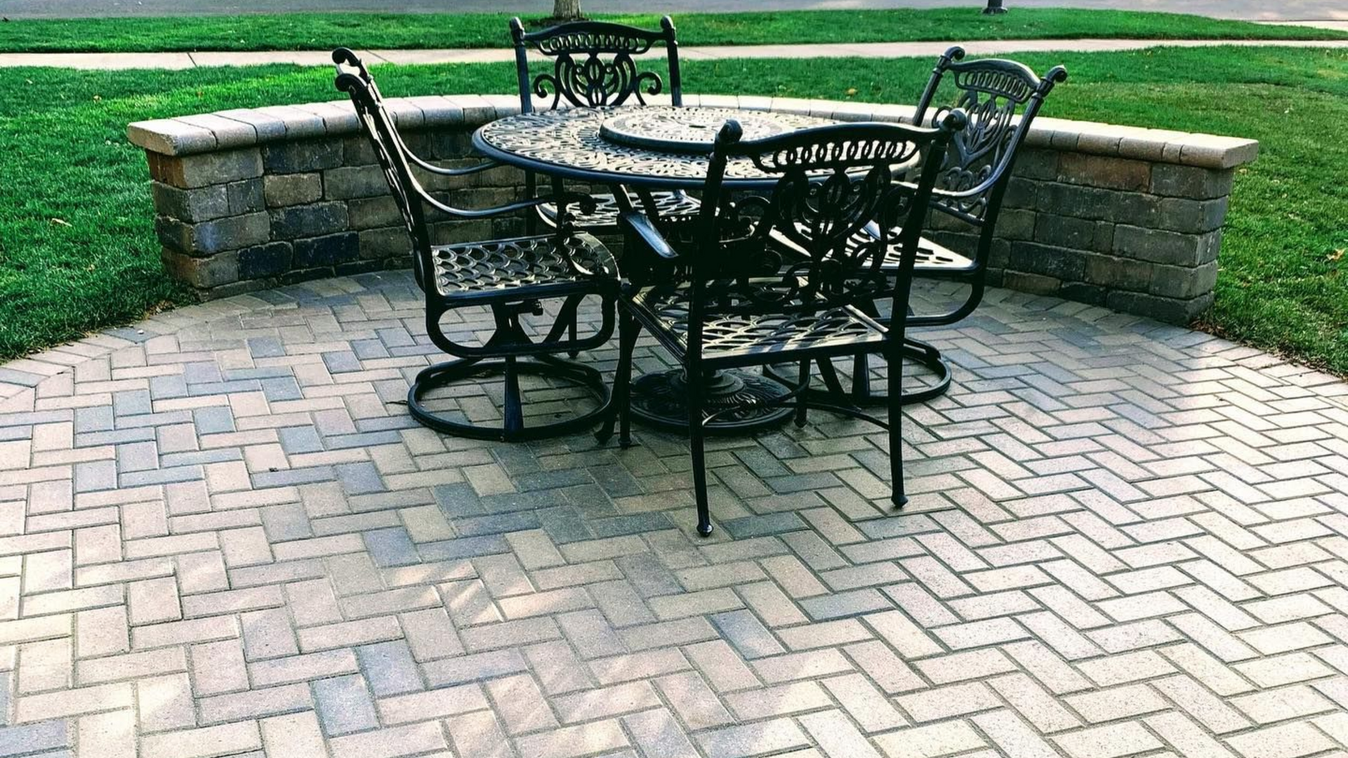 Patio with black metal table and chairs, brick pattern pavers, small stone wall, and green grass.