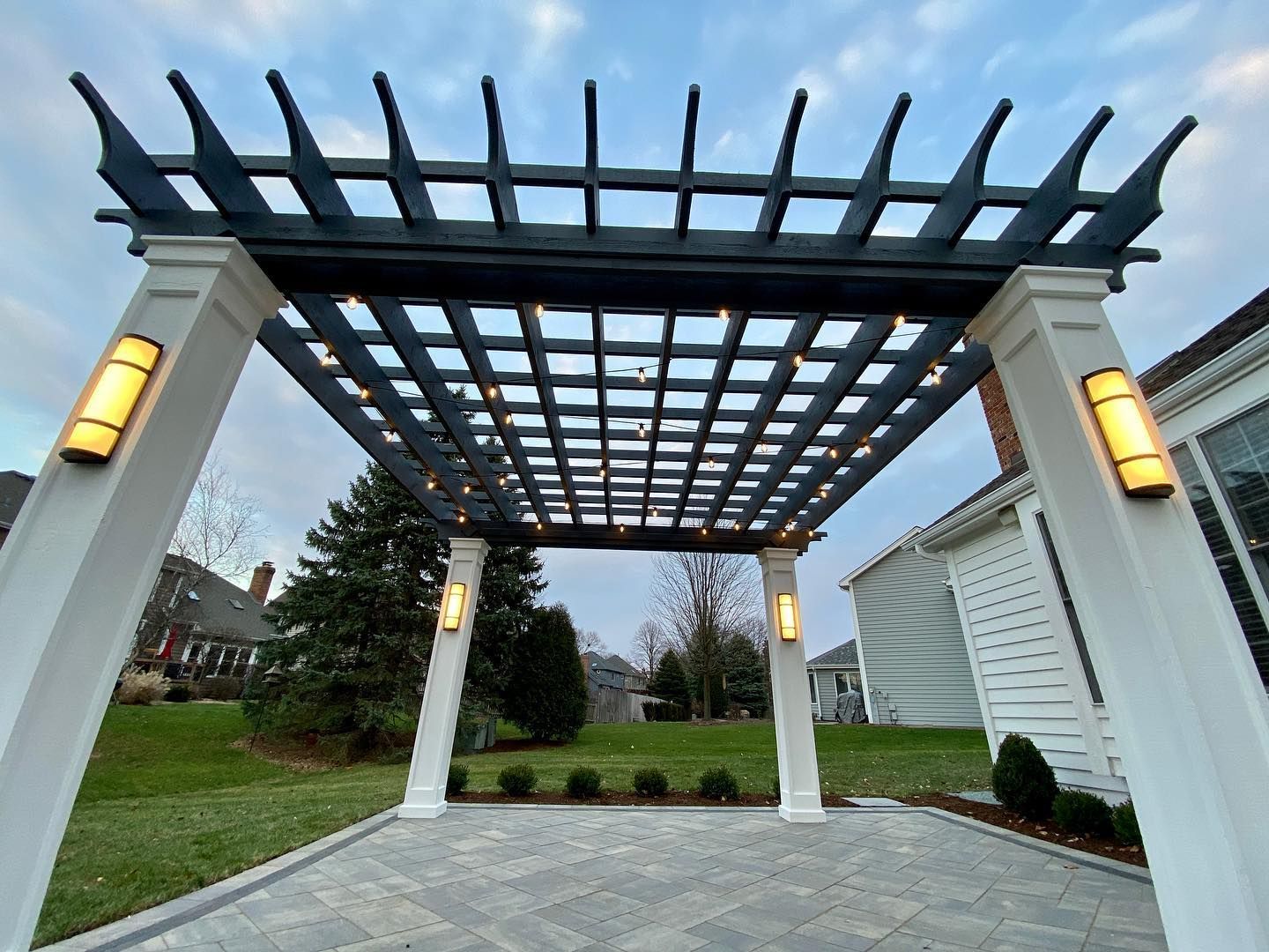 Pergola with string lights over a patio, lit by rectangular wall sconces, surrounded by lawn and a house.