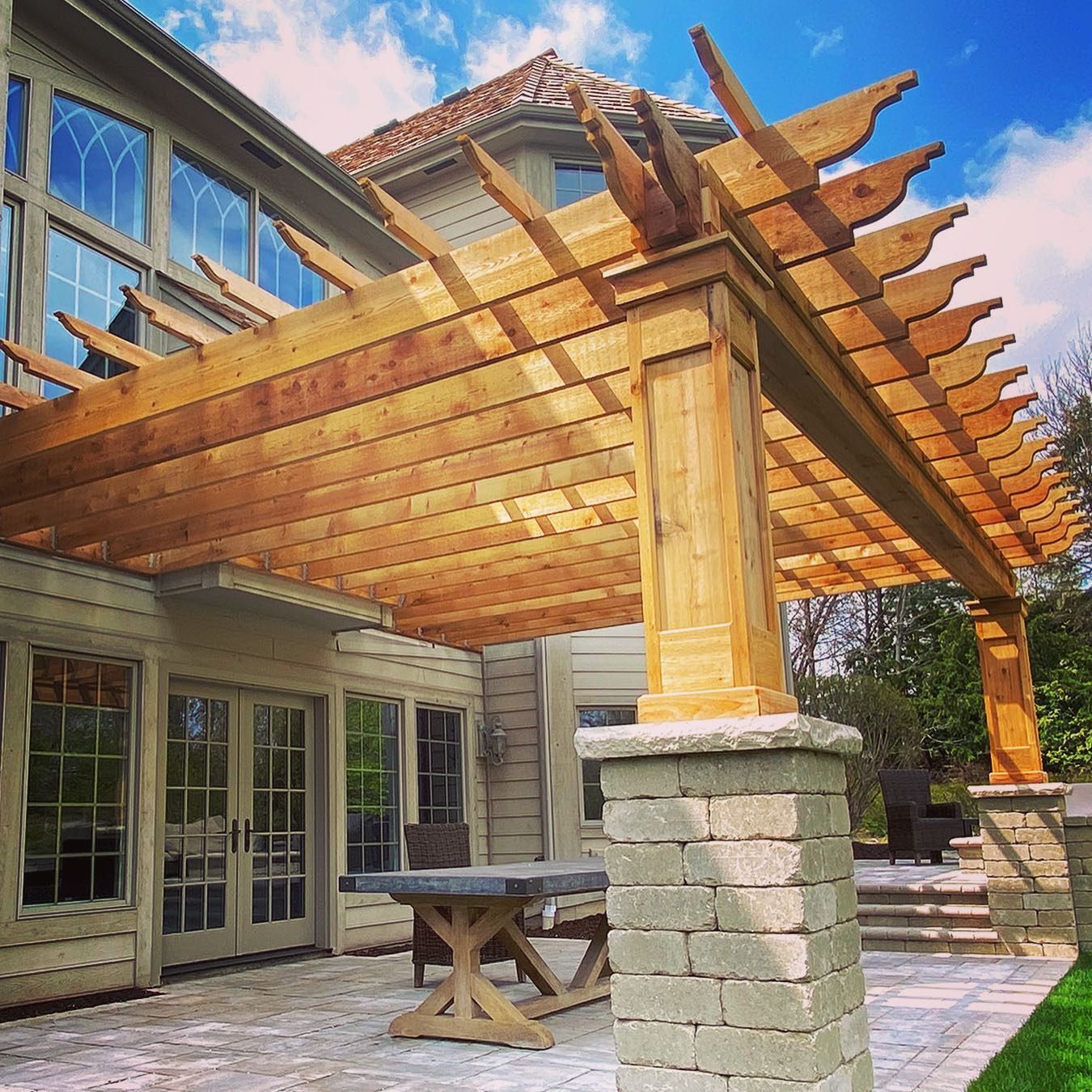 Wooden pergola over a patio with a table and chairs, attached to a house with large windows.