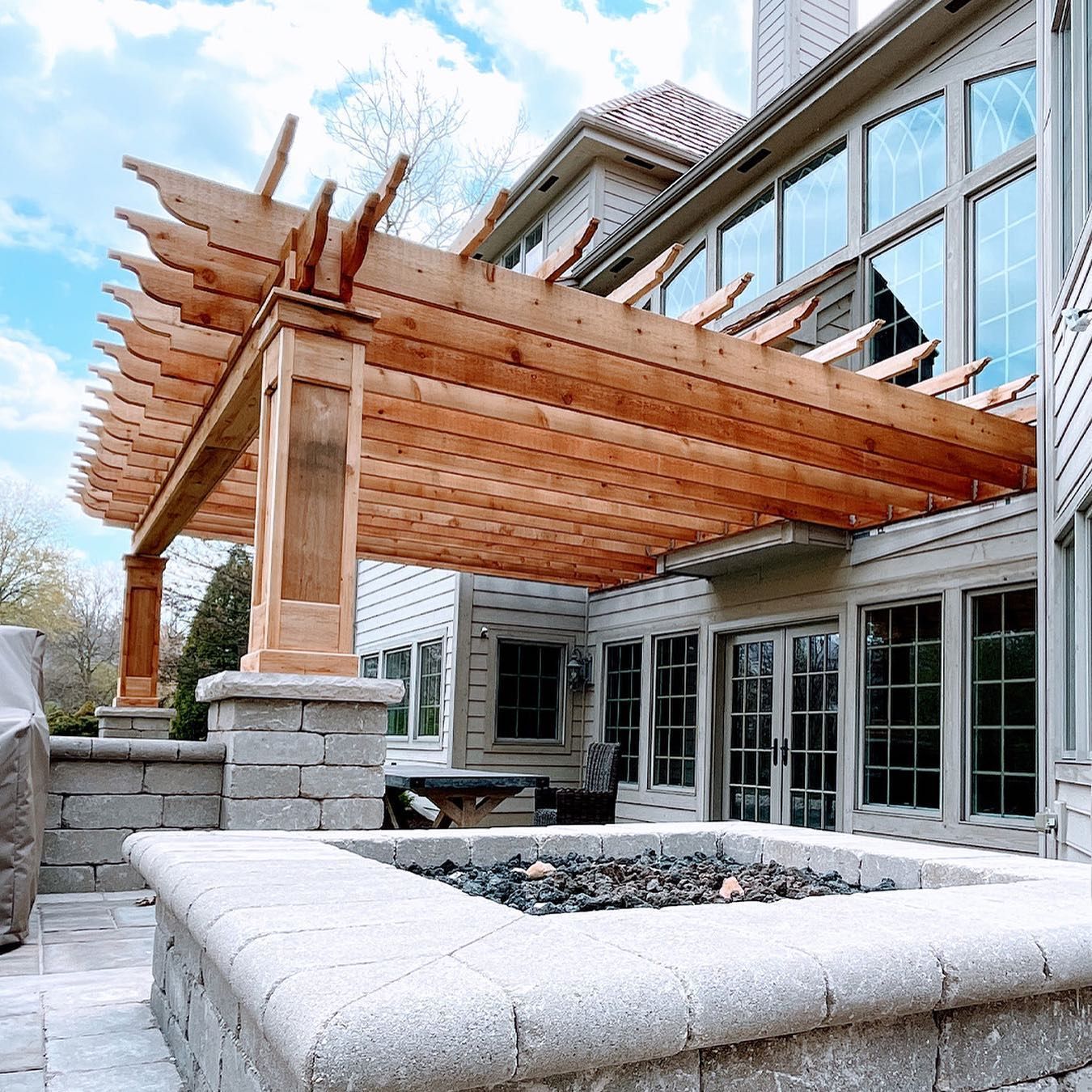 Pergola over patio with a fire pit, next to a two-story house with large windows.
