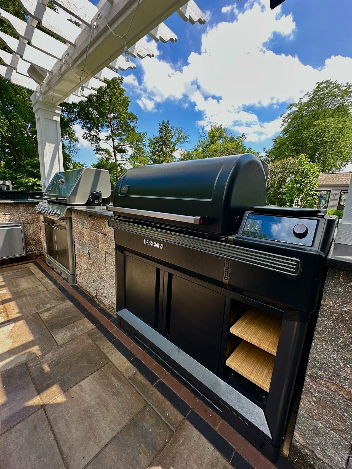 Outdoor built-in grill station with a black grill, digital display, and storage cabinet, under a white pergola on a stone patio.