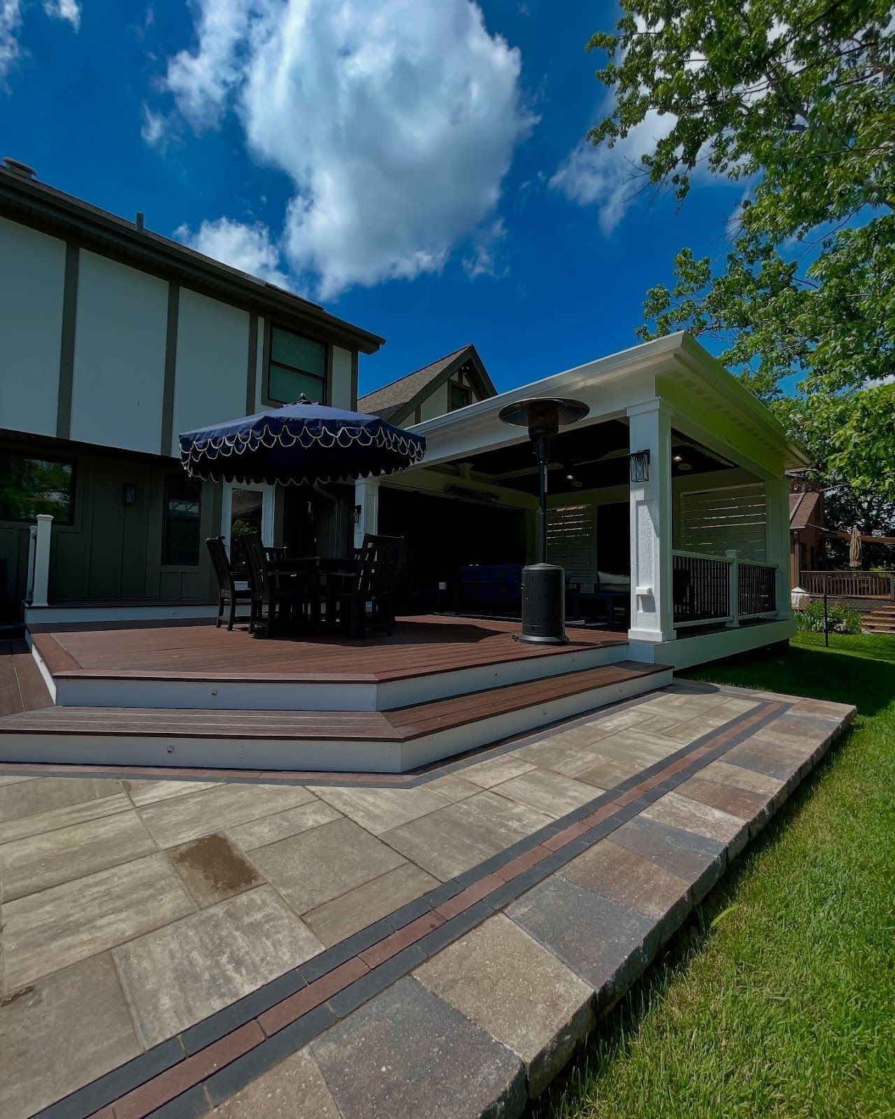 Patio with deck, pavers, umbrella, and covered seating area. House in background under blue sky.