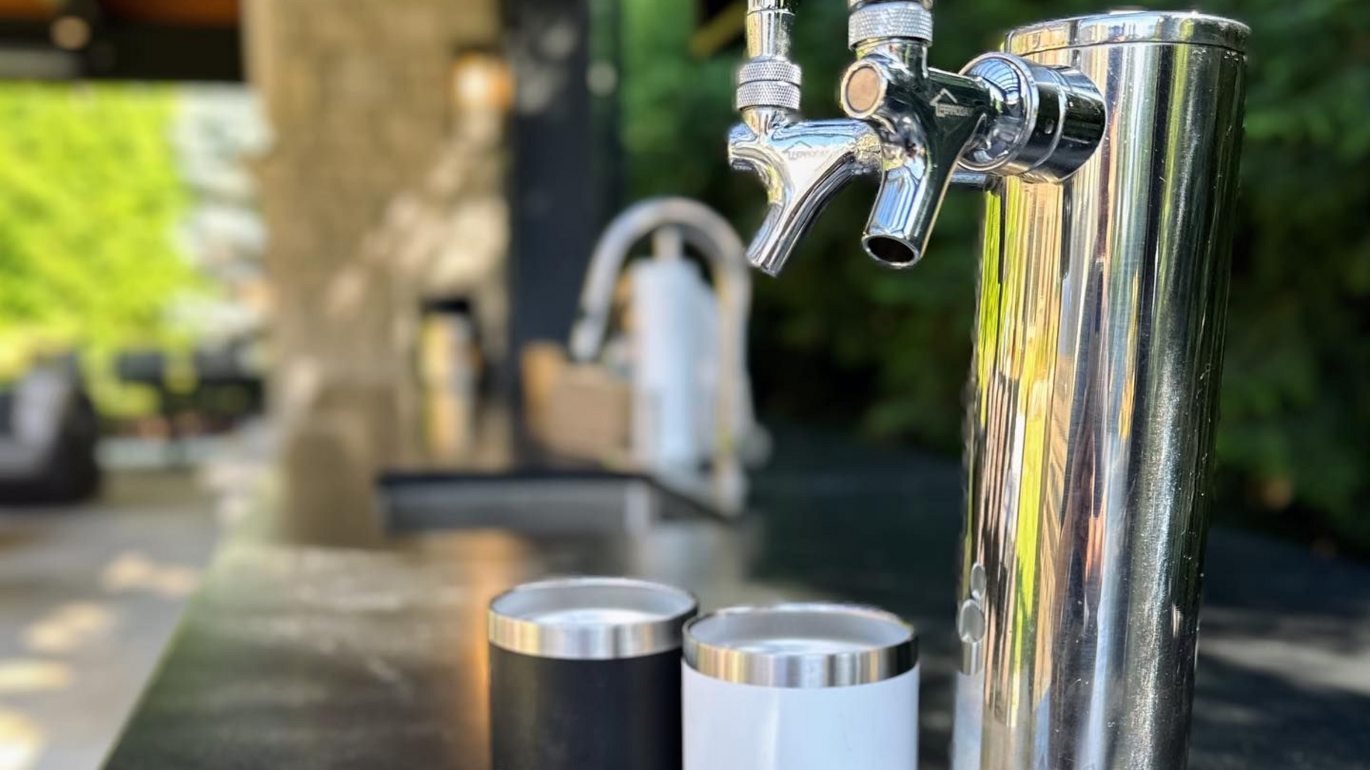 Chrome beer tap with two stainless steel cups on outdoor bar.