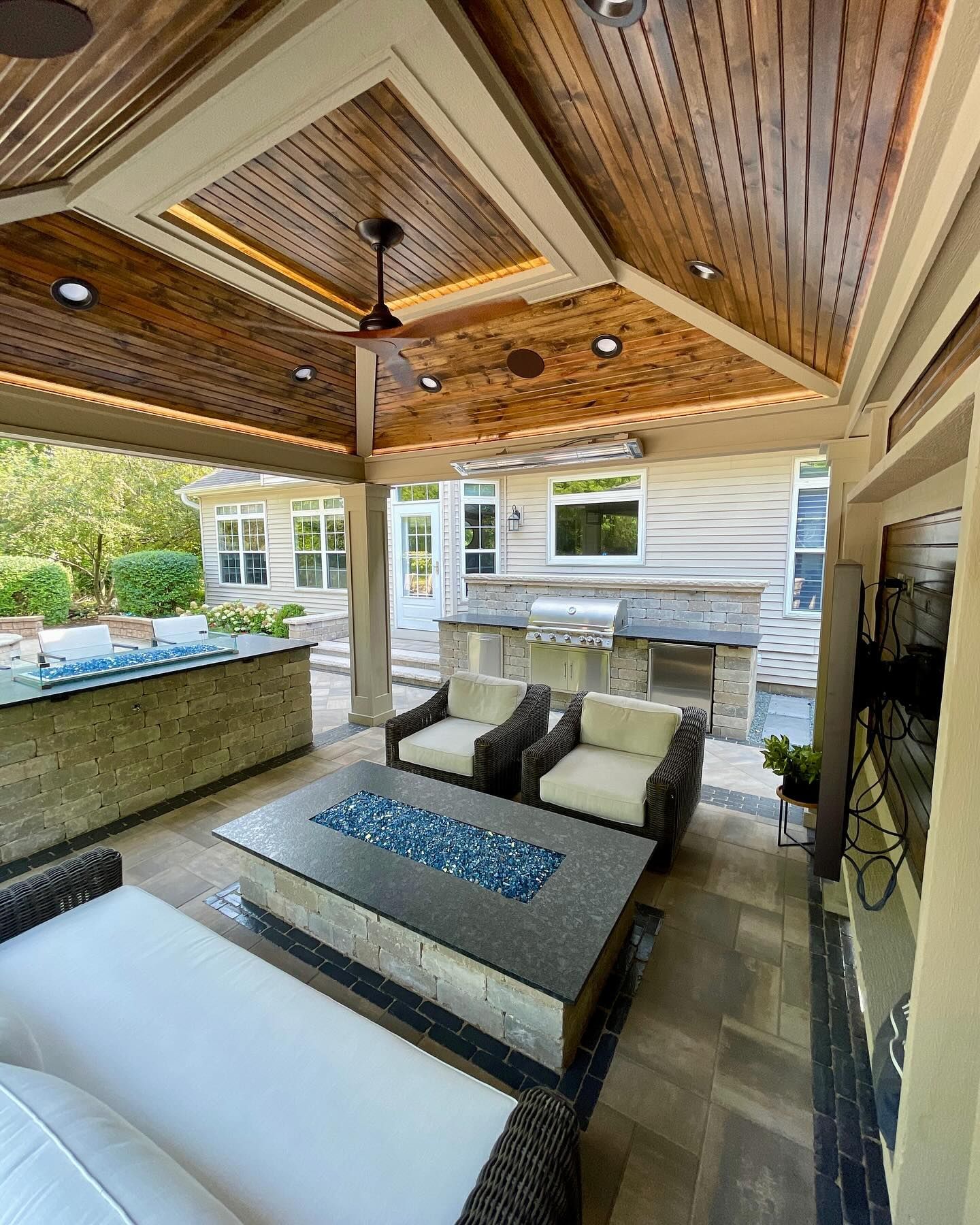 Outdoor covered patio with seating, fire pit, and built-in kitchen. Wooden ceiling and stone accents.