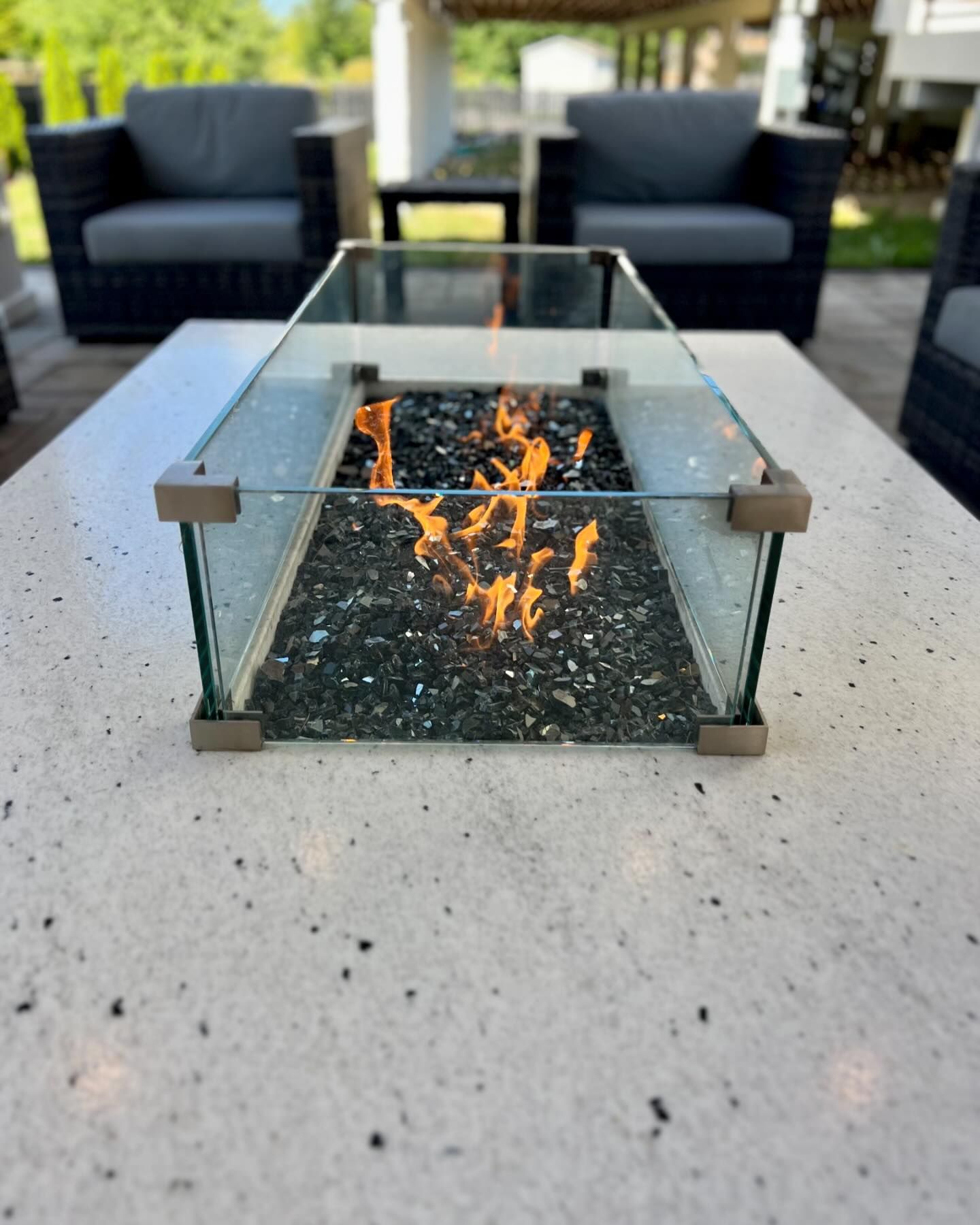 Fire pit on a white speckled table, glass sides, flames, surrounded by chairs.