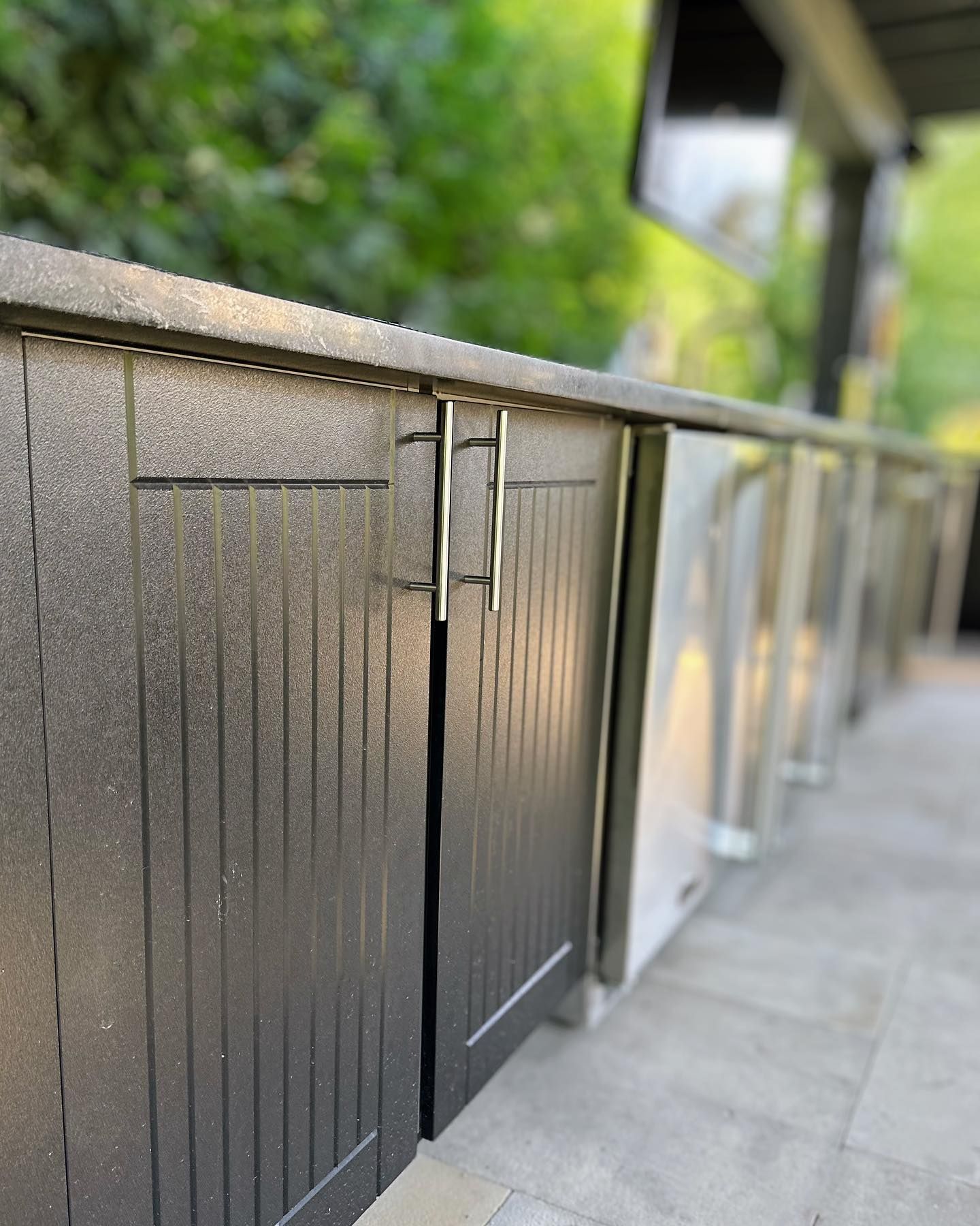 Outdoor kitchen cabinets, textured black, with silver handles. Stainless steel appliances in the background, green foliage.