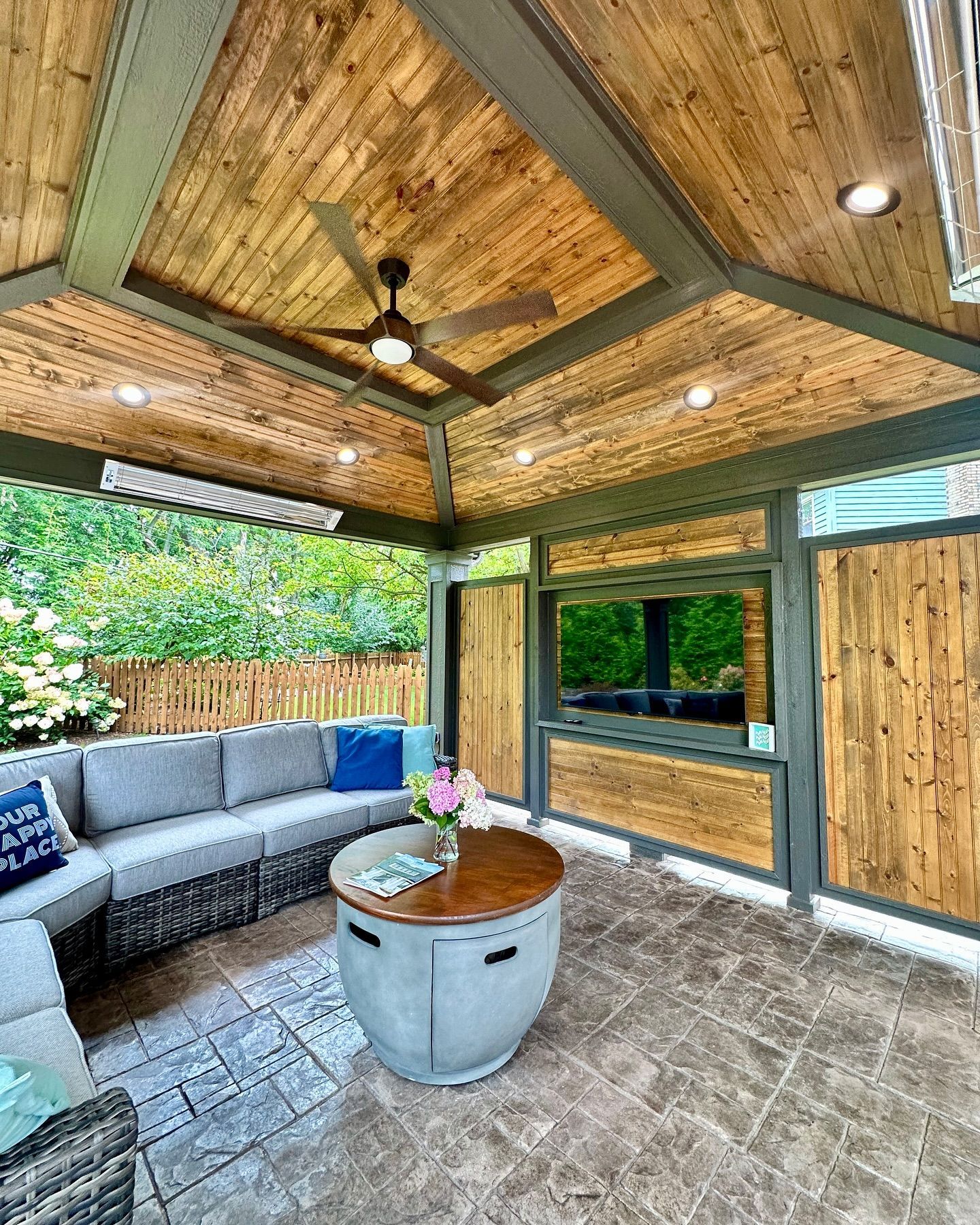 Outdoor patio with sectional sofa, wood ceiling, round fire pit table, and large-screen television.