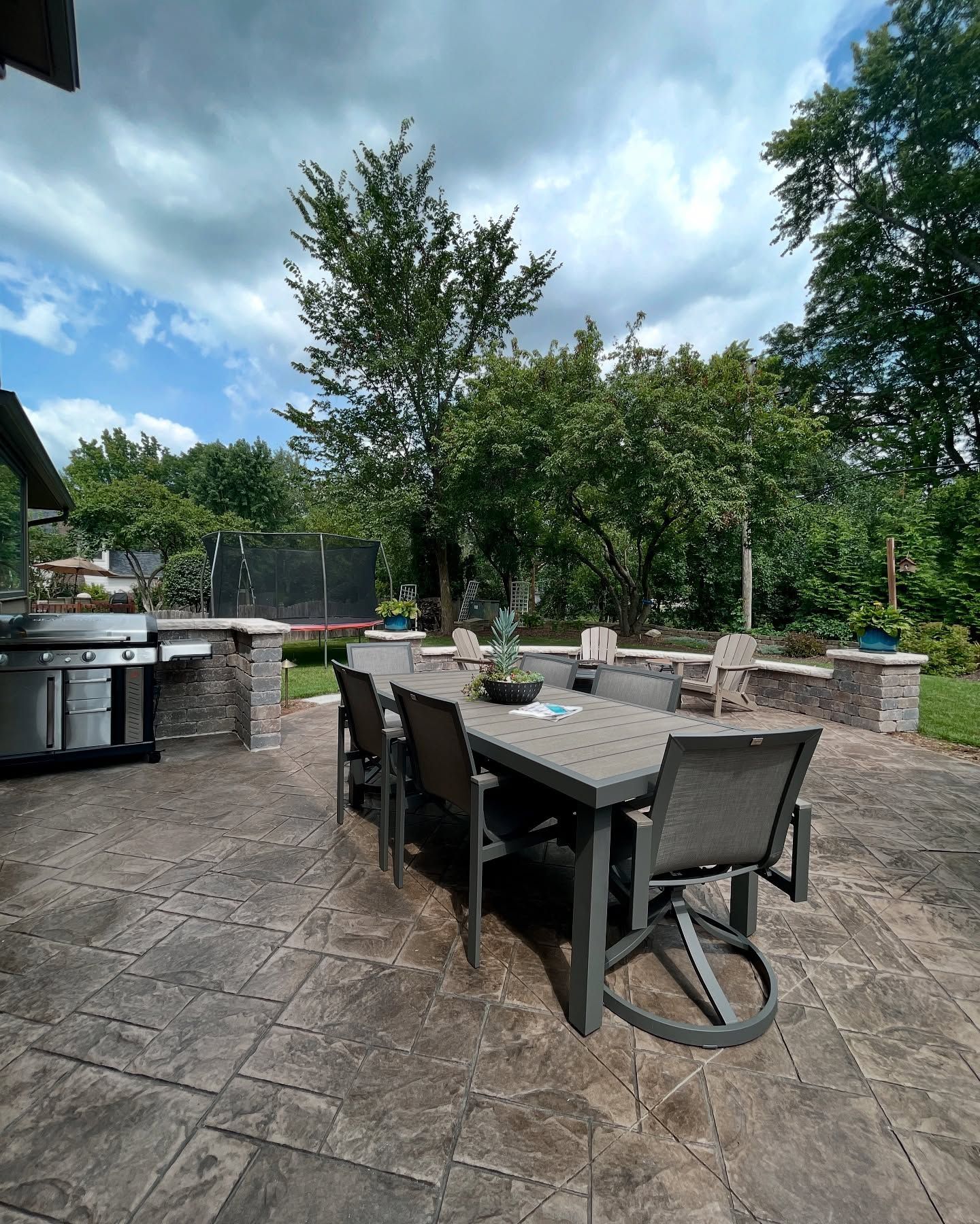 Patio with outdoor dining table, grill, and trees under cloudy sky.